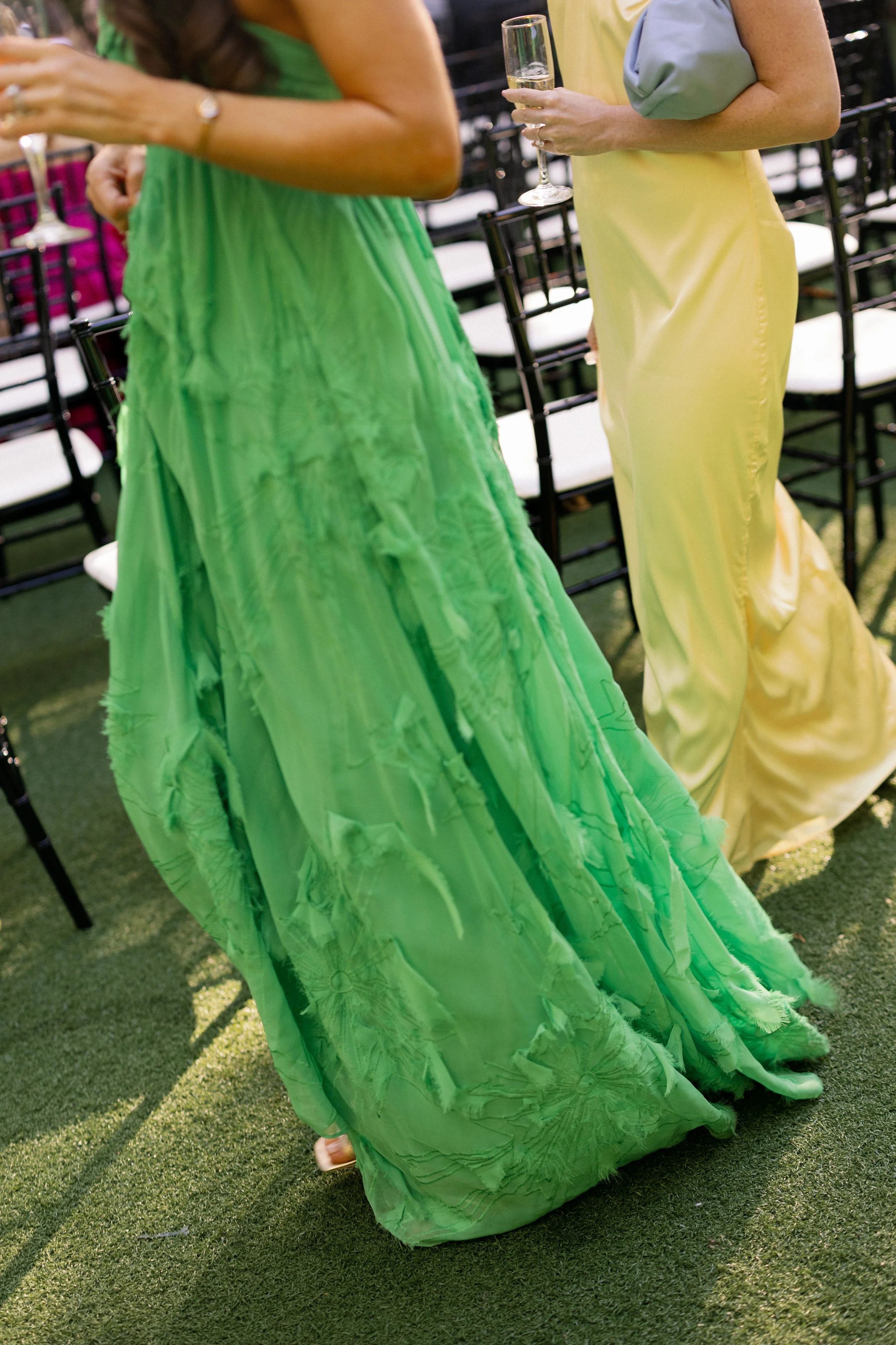 Two women wearing colorful dresses, one in a bright green gown and the other in yellow, are standing outside on a grassy area at a social gathering, holding champagne glasses.