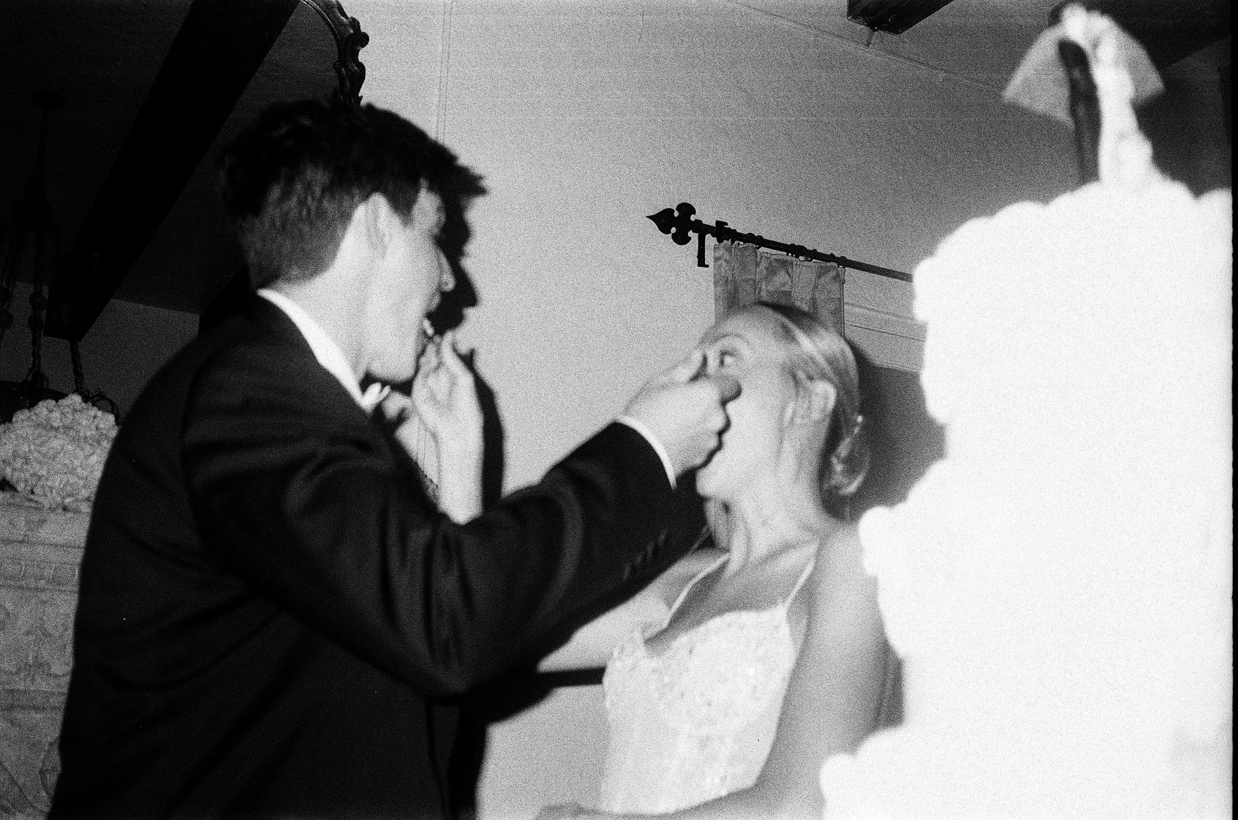 A person in a tuxedo is placing a ring on the finger of a woman in a wedding dress, both standing in front of a wedding cake. The woman is looking at the person, and the scene appears to be part of a wedding ceremony or celebration.