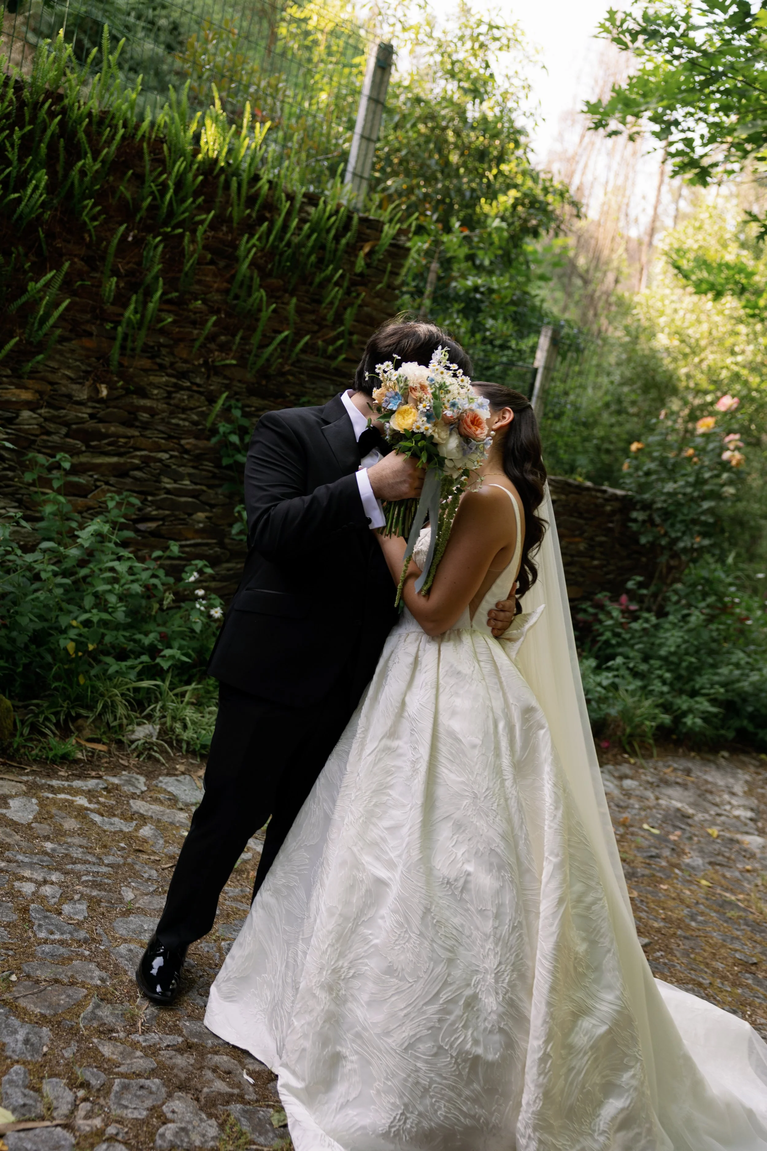 A bride and groom sharing a kiss, with the groom holding a bouquet of flowers covering their faces, outdoors with greenery and a cobblestone pathway.