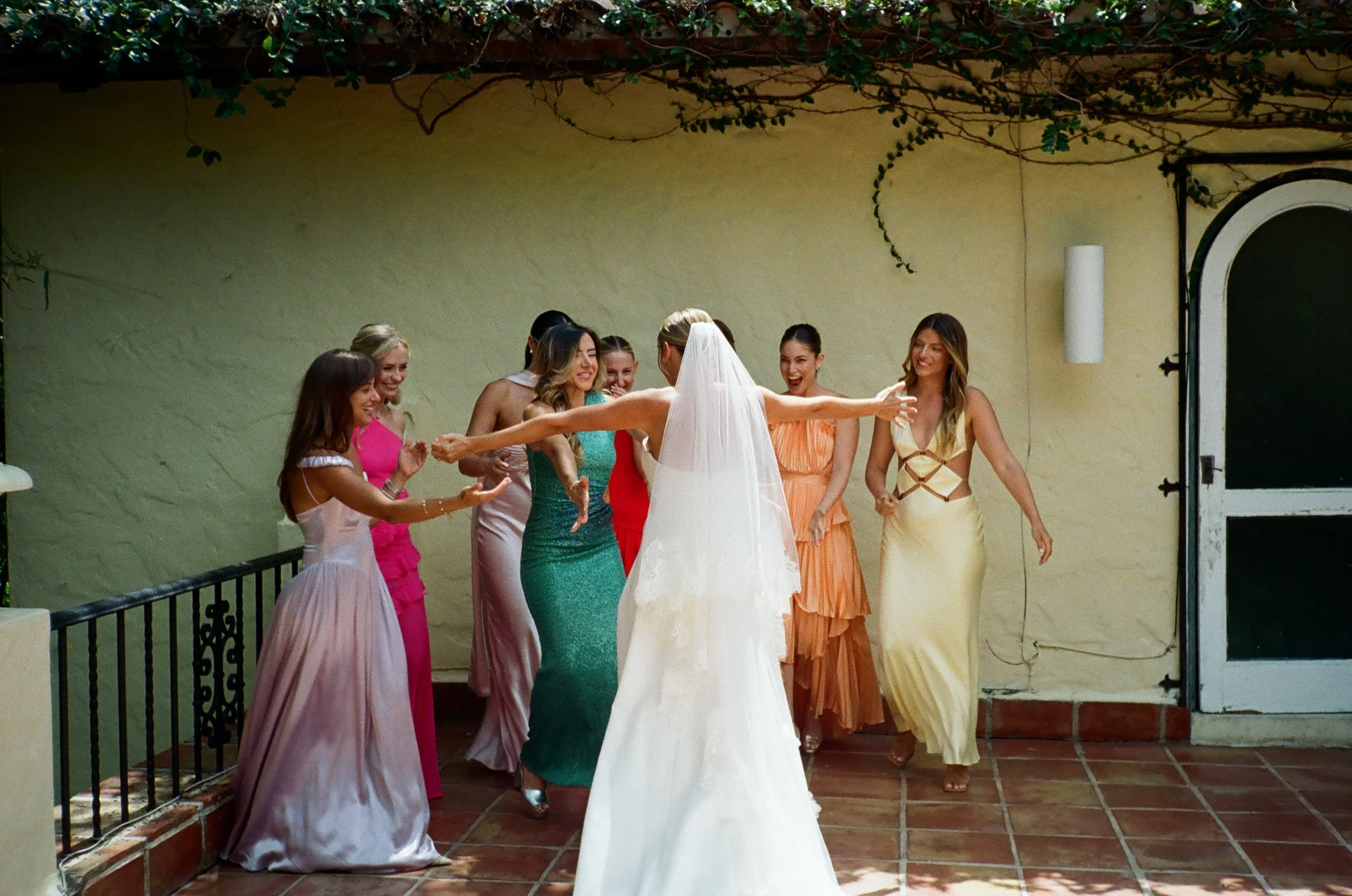 A bride in a white wedding dress and veil is surrounded by her bridesmaids, dressed in colorful gowns, as they laugh and celebrate outdoors under a vine-covered pergola during a wedding.
