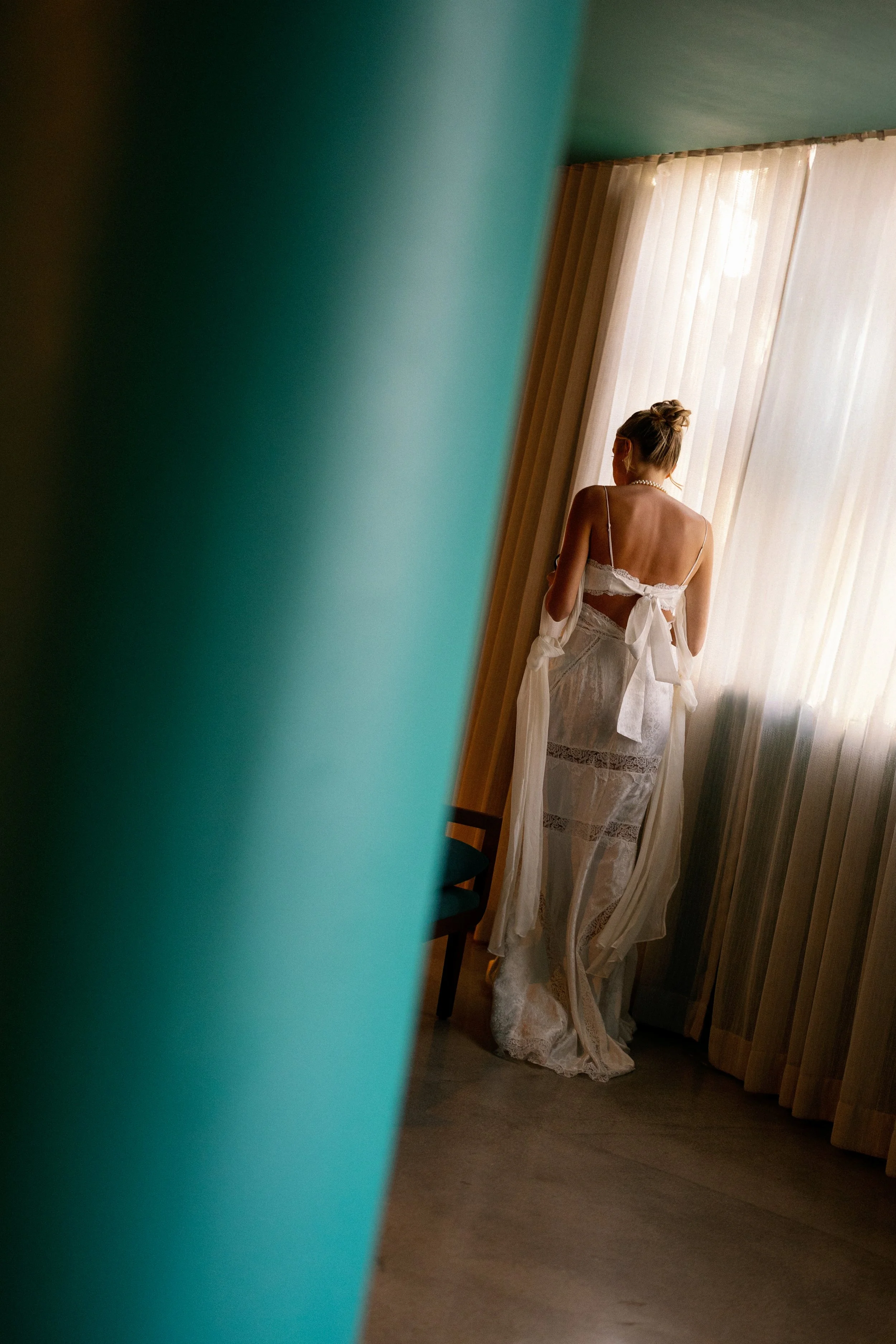 A woman dressed in a white lace dress, possibly a wedding gown, standing by a window with sheer curtains, captured through a partially opened door or curtain.