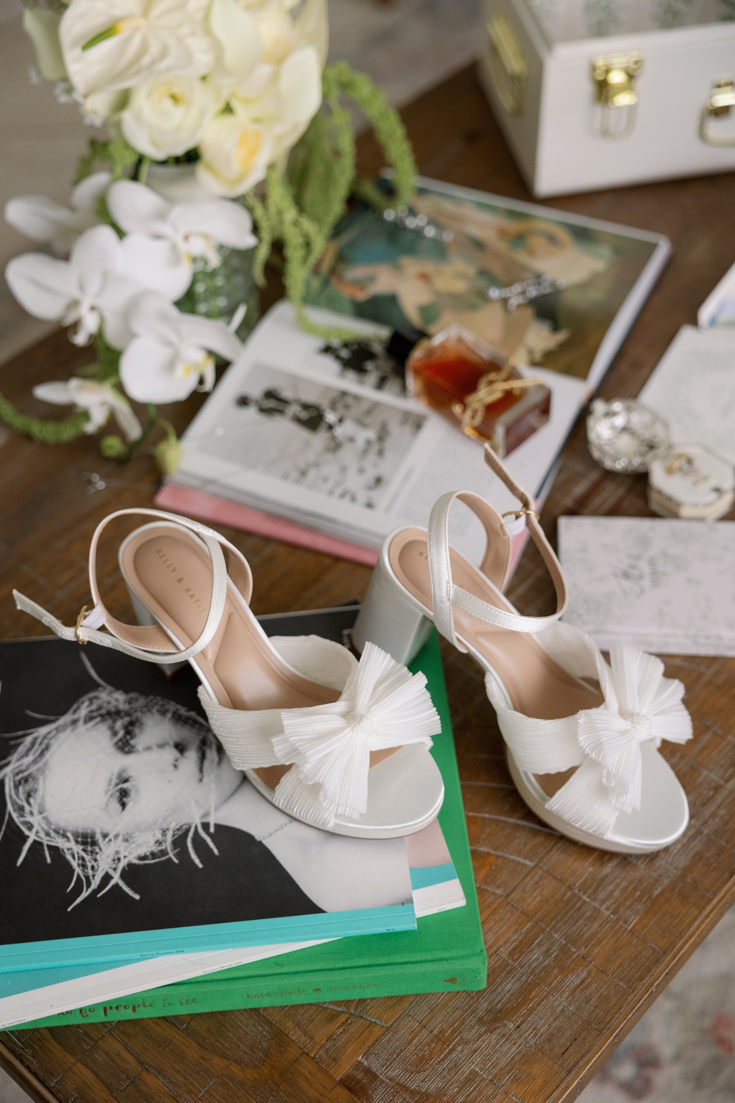 A pair of white high-heeled shoes with large bows on the front, placed on top of books and magazines on a wooden table. There is a bouquet of white flowers and some decorative objects nearby.