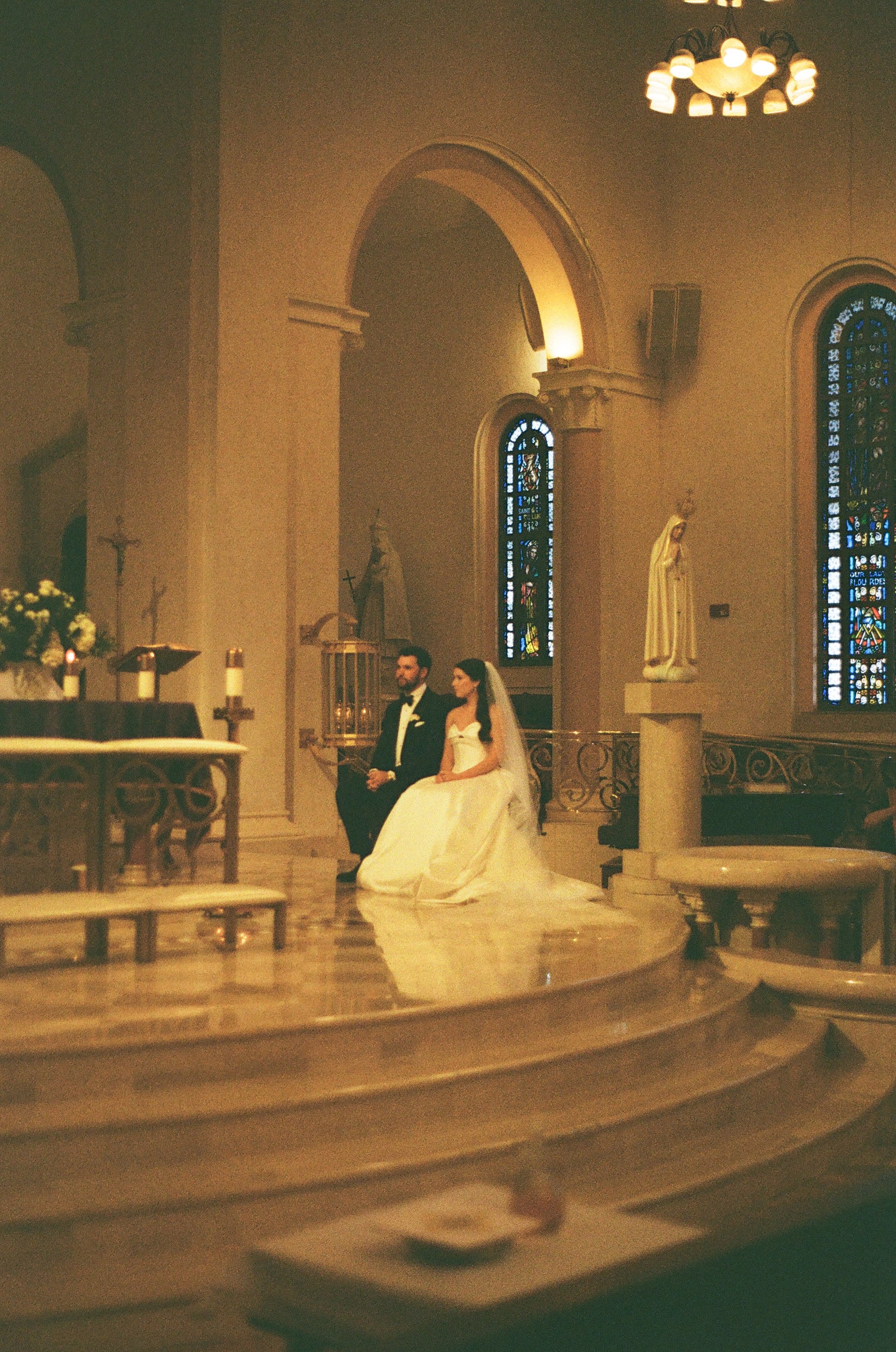 A bride and groom standing together inside a church during a wedding ceremony. The church has stained glass windows, religious statues, and a chandelier hanging from the ceiling. The altar is decorated with flowers and candles.