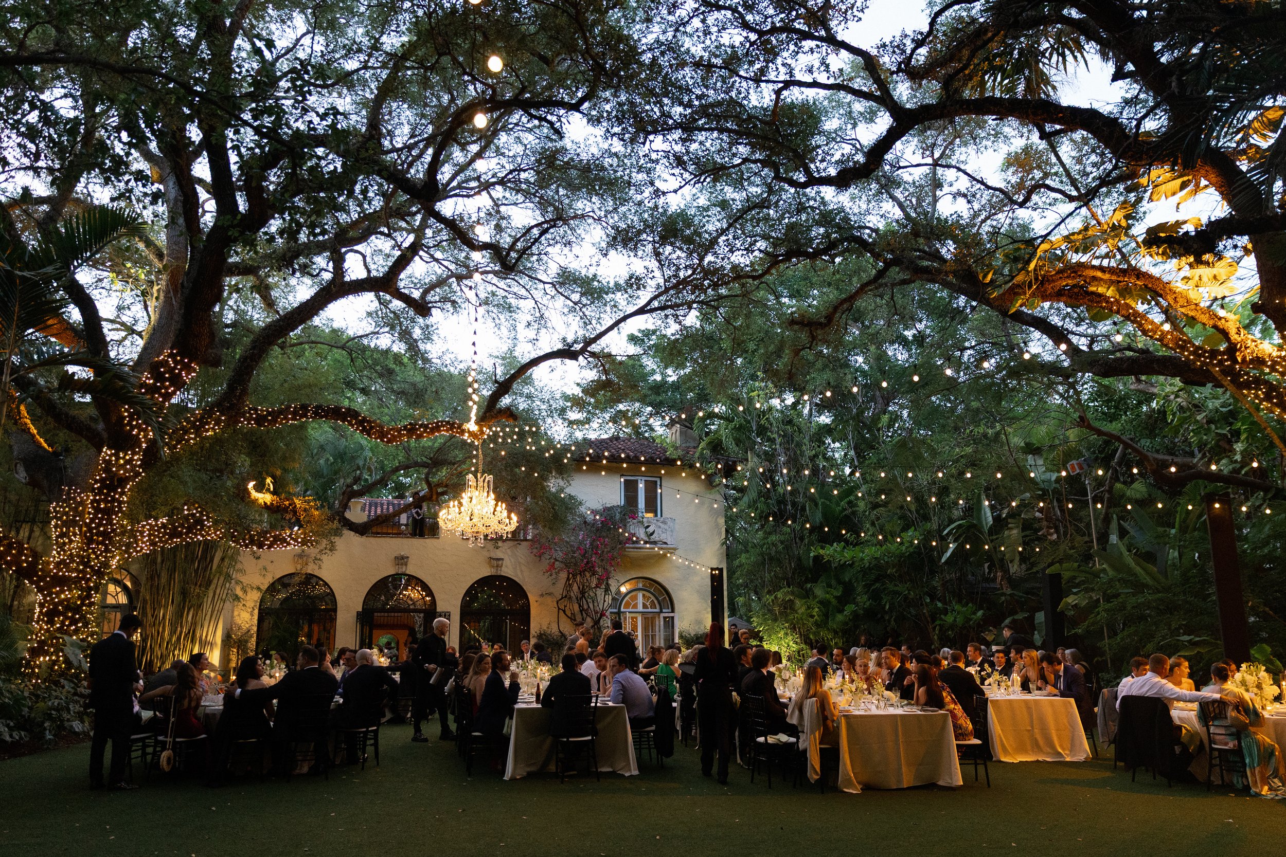 Outdoor evening celebration with large gathering of people seated at tables under string lights and chandeliers amid trees decorated with fairy lights, next to a yellow building with arched windows.