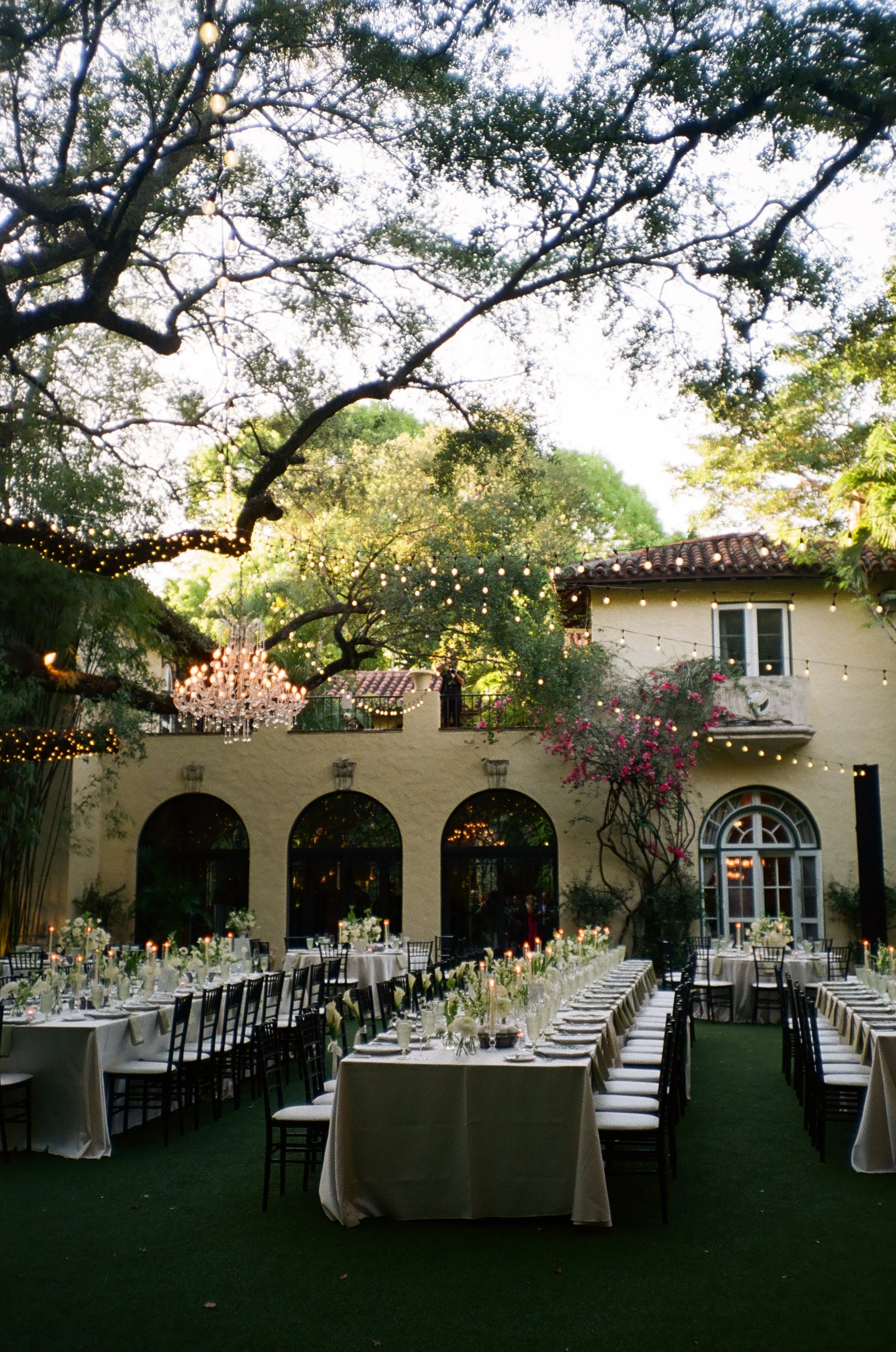 Outdoor wedding reception set up with long banquet tables, chairs, floral centerpieces, and string lights hanging from large trees on a garden patio.