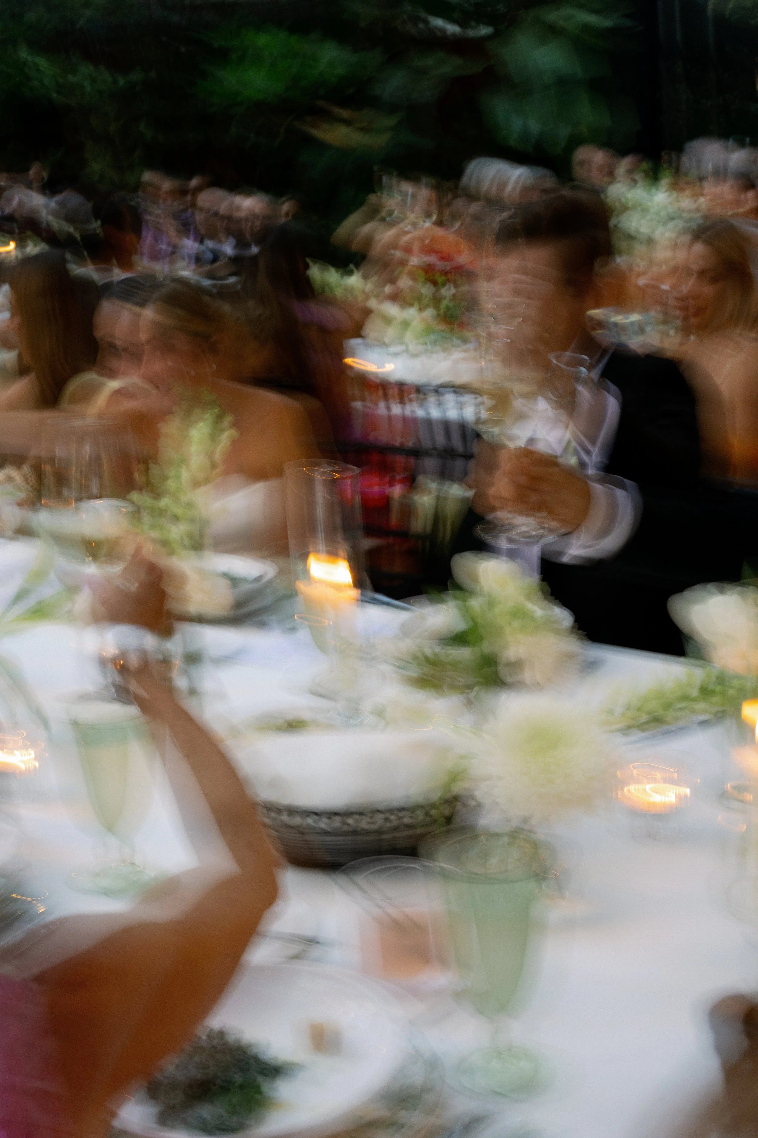 Blurred image of people at a formal event, sitting at a long table with white tablecloths, candles, and floral centerpieces, with a dark outdoor background.