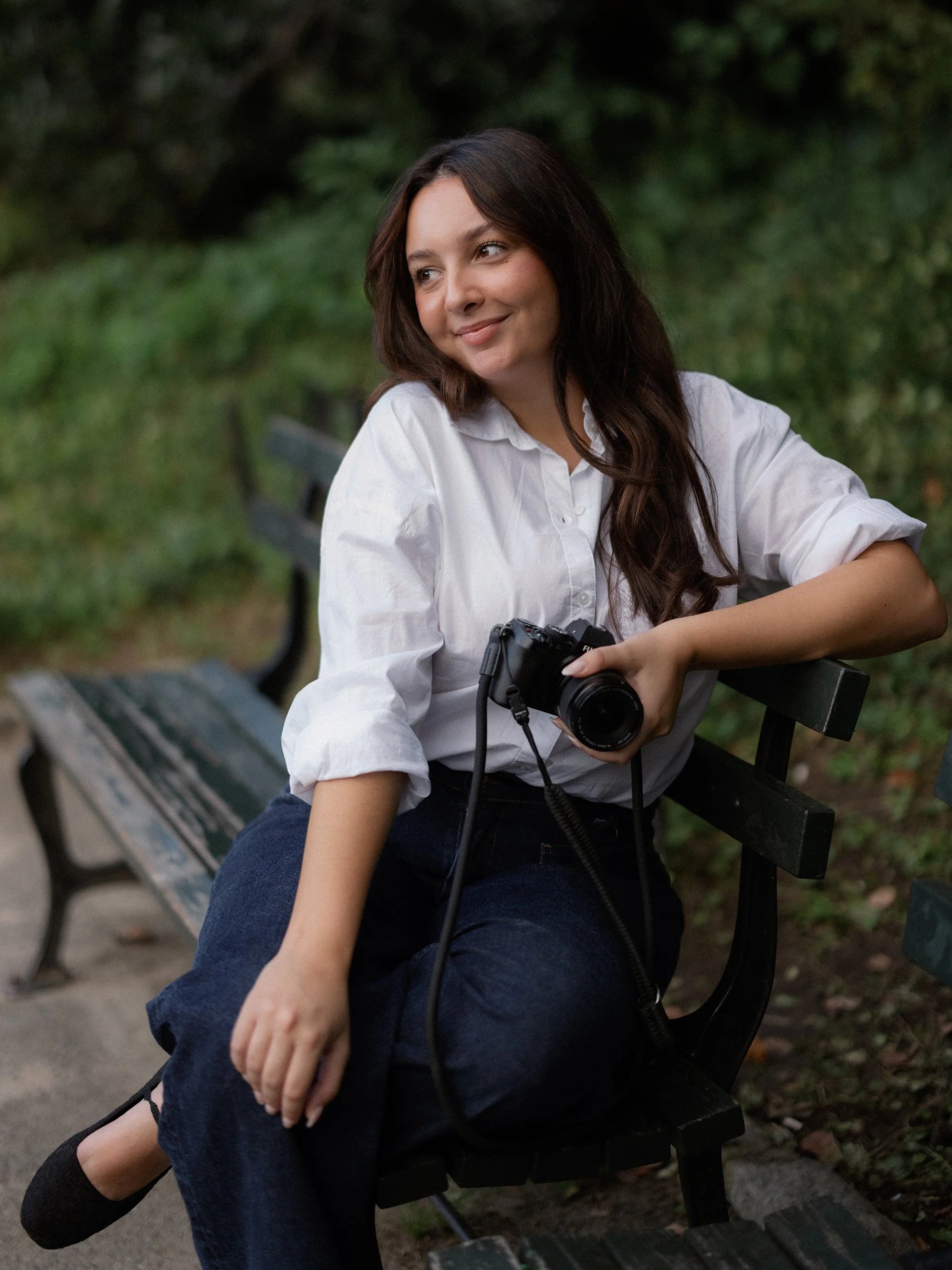 Young woman with long brown hair sitting on a park bench holding a camera, smiling softly, wearing a white shirt and dark blue pants, with greenery in the background.