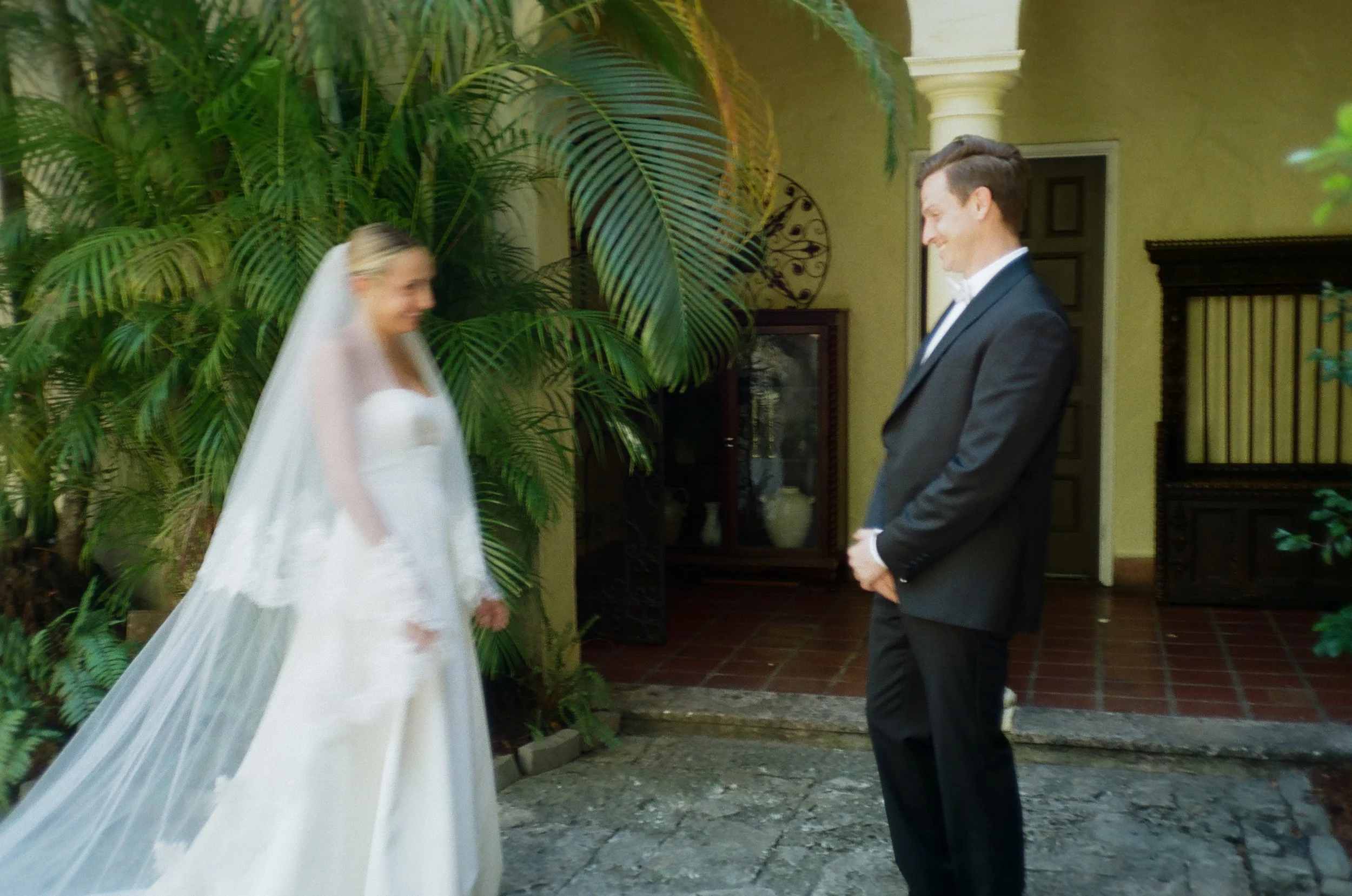 A bride and groom stand facing each other indoors, with greenery and decorative elements in the background. The bride is wearing a white wedding gown and veil, while the groom is dressed in a dark suit with a white shirt and bow tie. They appear to b