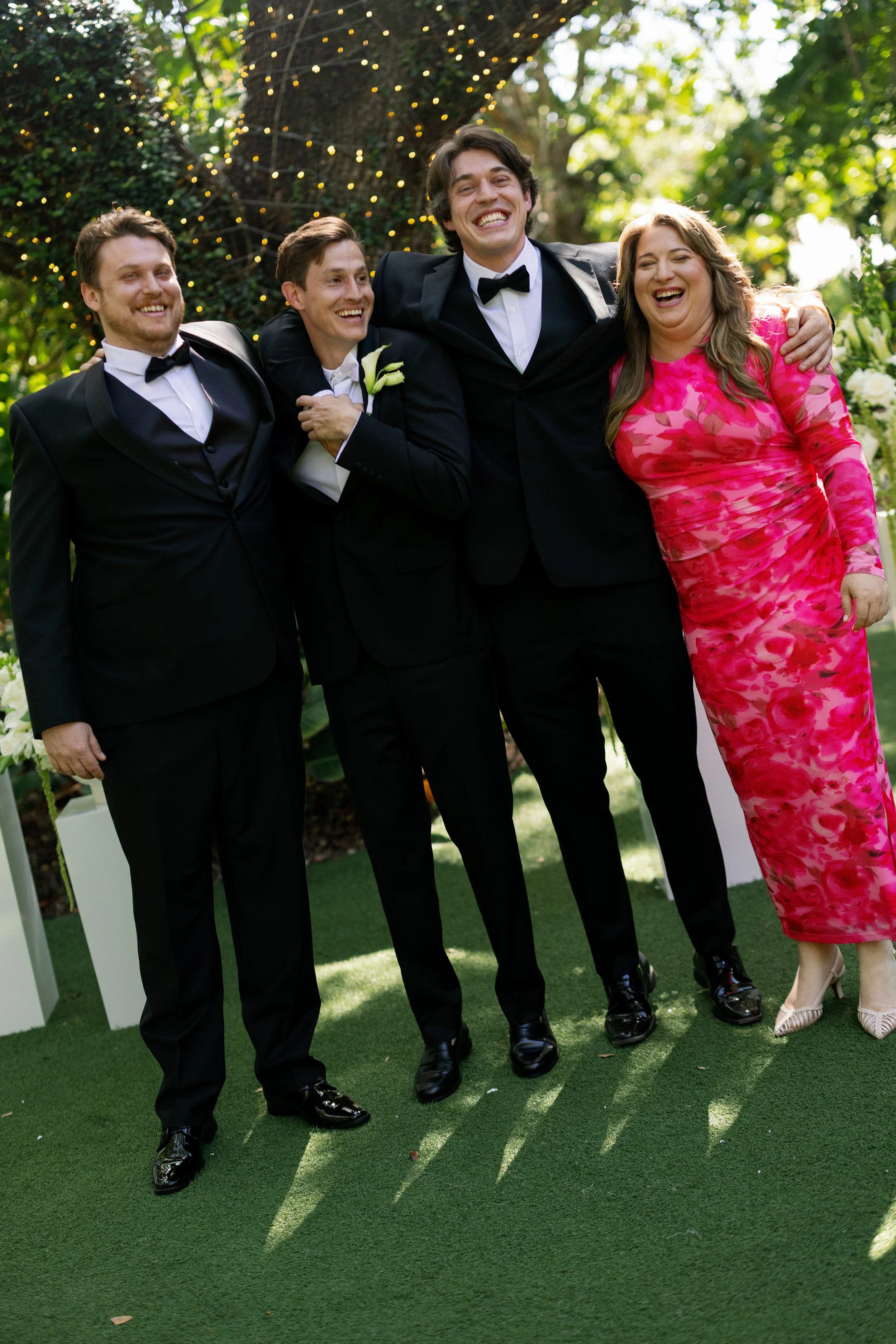 Group of five friends celebrating at a wedding outdoors, dressed in formal attire, with smiles, standing on green grass with trees and fairy lights in the background.