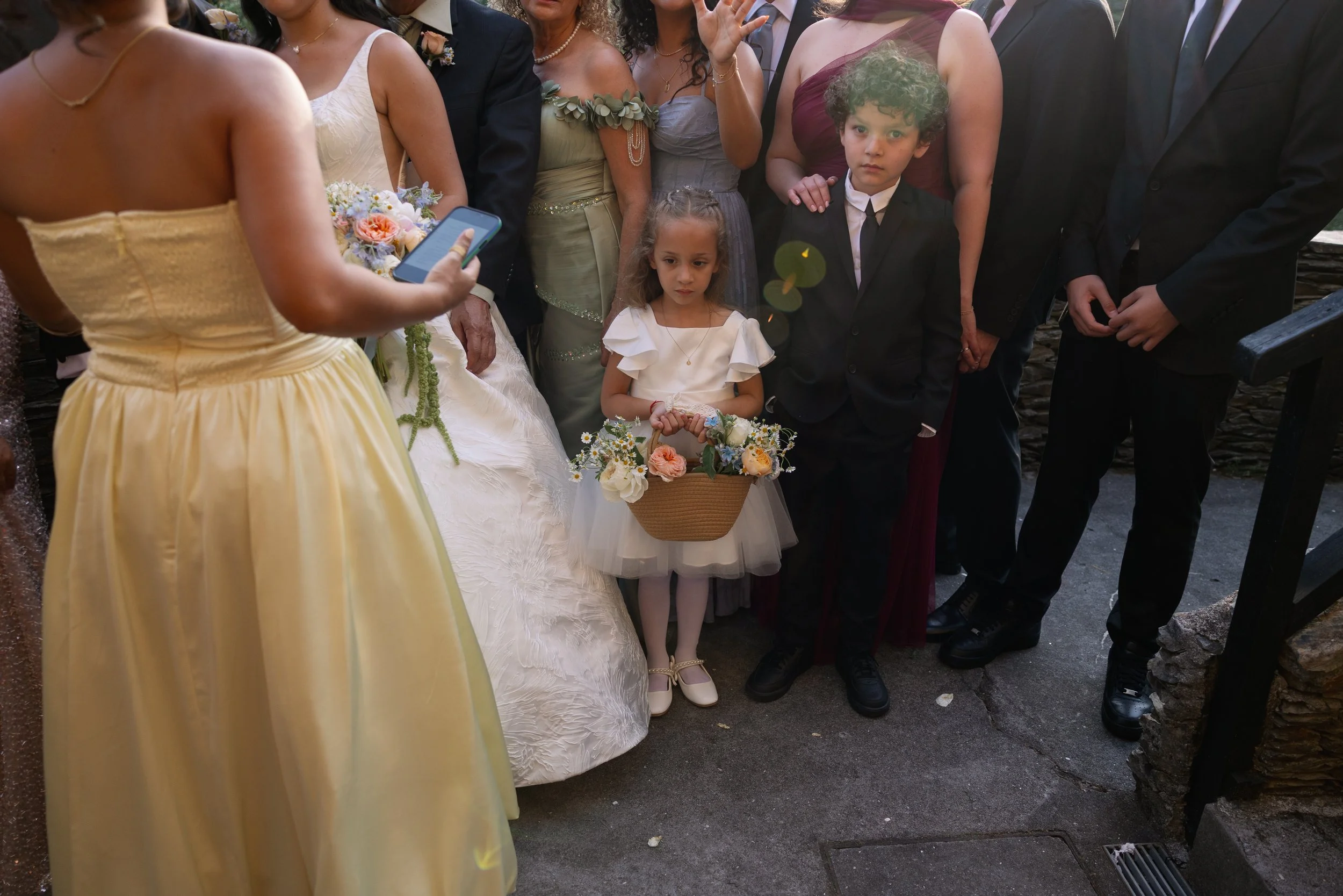 A group of formally dressed people, including young children, gathered outdoors for a wedding ceremony. The bride, dressed in white, is visible with her back to the camera, holding a bouquet and a phone. Two young children, a girl in a white dress with ruffles holding a small basket of flowers, and a boy in a black suit, are standing in the foreground. The scene captures a moment during the wedding celebration.