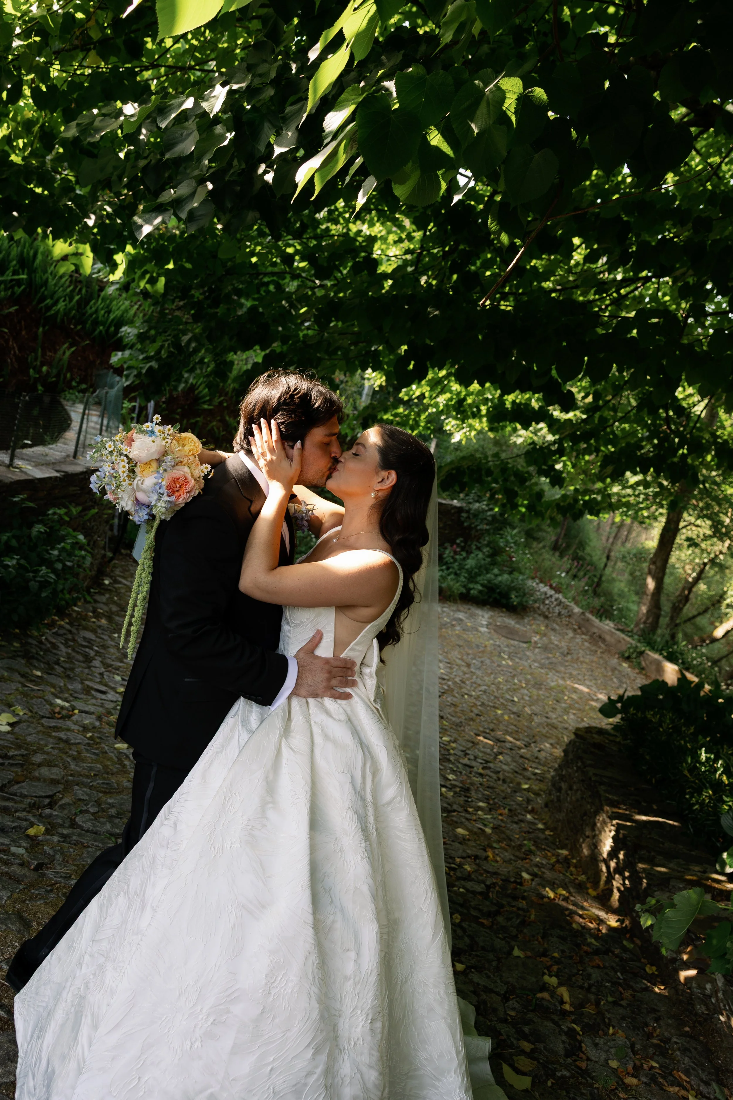 A bride and groom sharing a kiss outdoors under a tree with lush green leaves, the bride wearing a white wedding gown and holding a bouquet of colorful flowers, and the groom dressed in a black tuxedo.