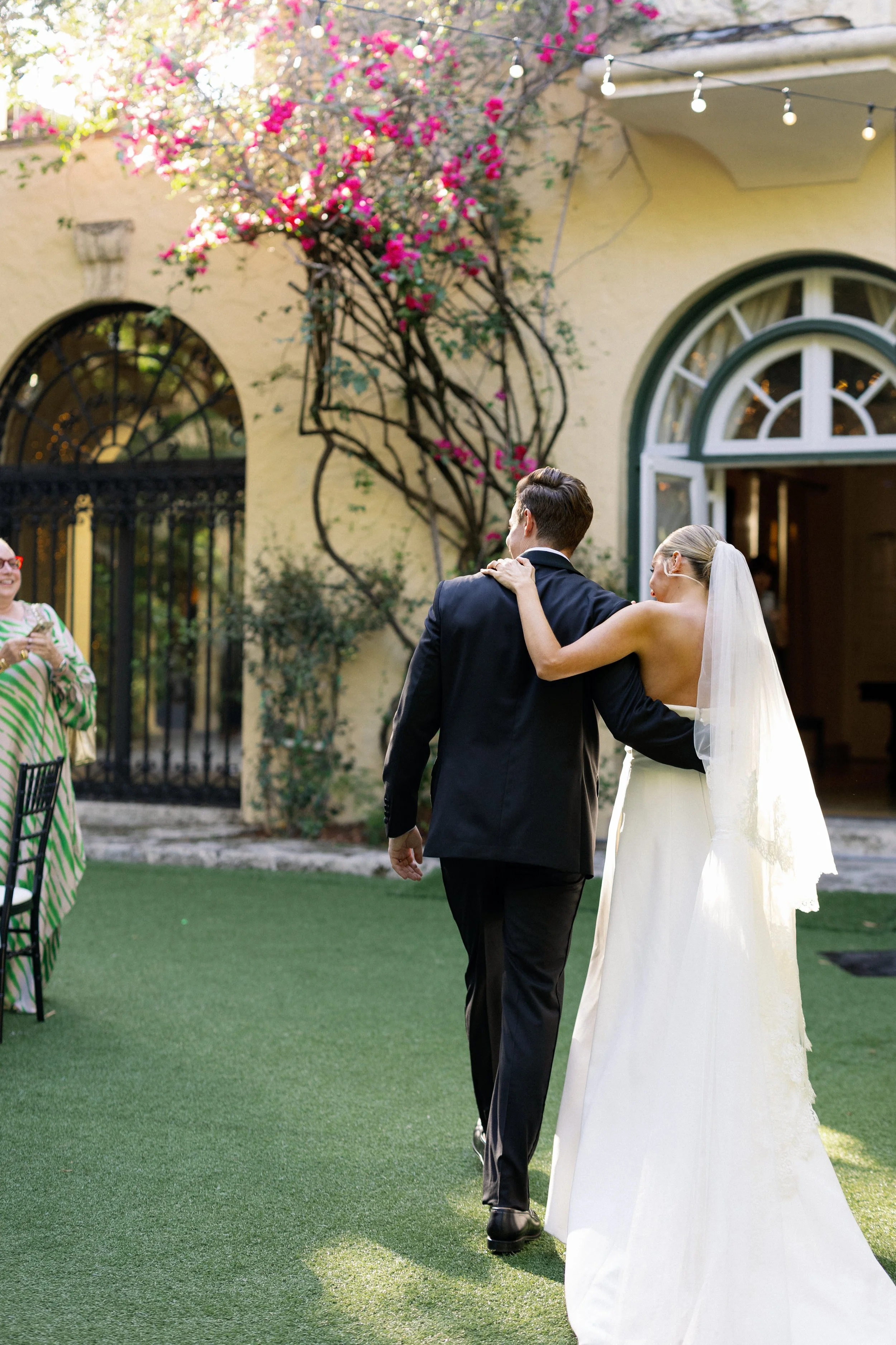 A bride and groom walking arm-in-arm outdoors during a wedding celebration. The bride is wearing a white wedding gown with a veil, and the groom is dressed in a dark suit. There are string lights overhead, a building with arched windows, and pink flo