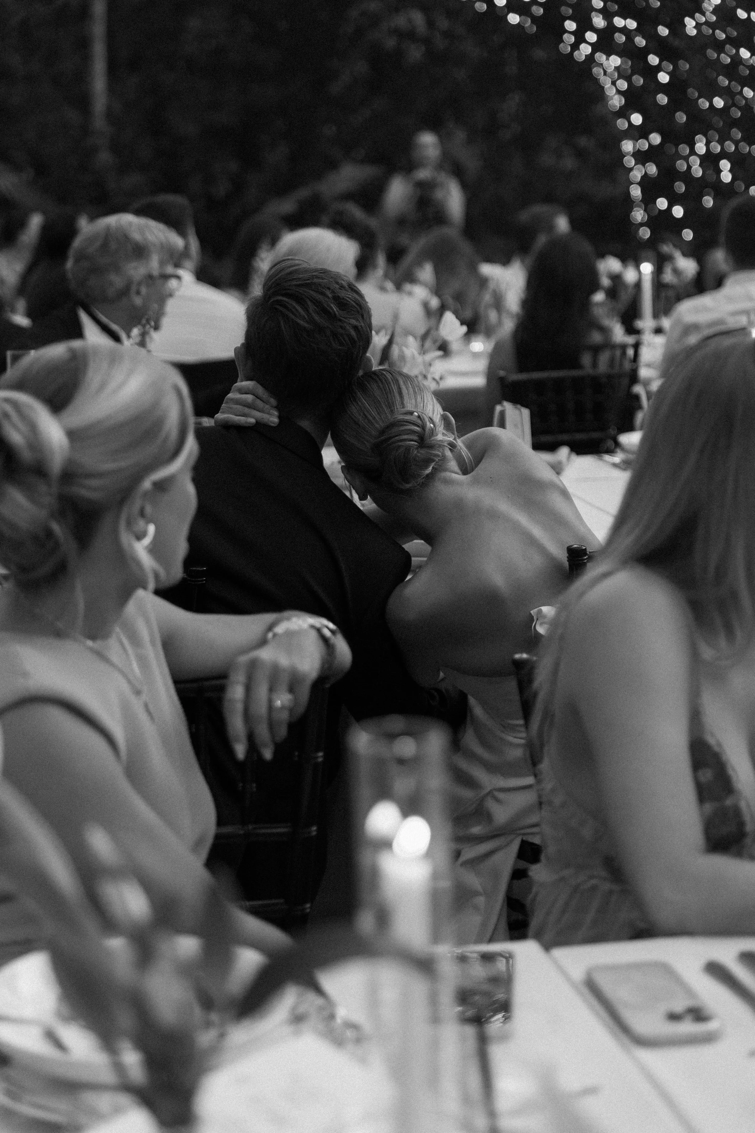 People sitting at a table outdoors during an evening event, with string lights in the background.