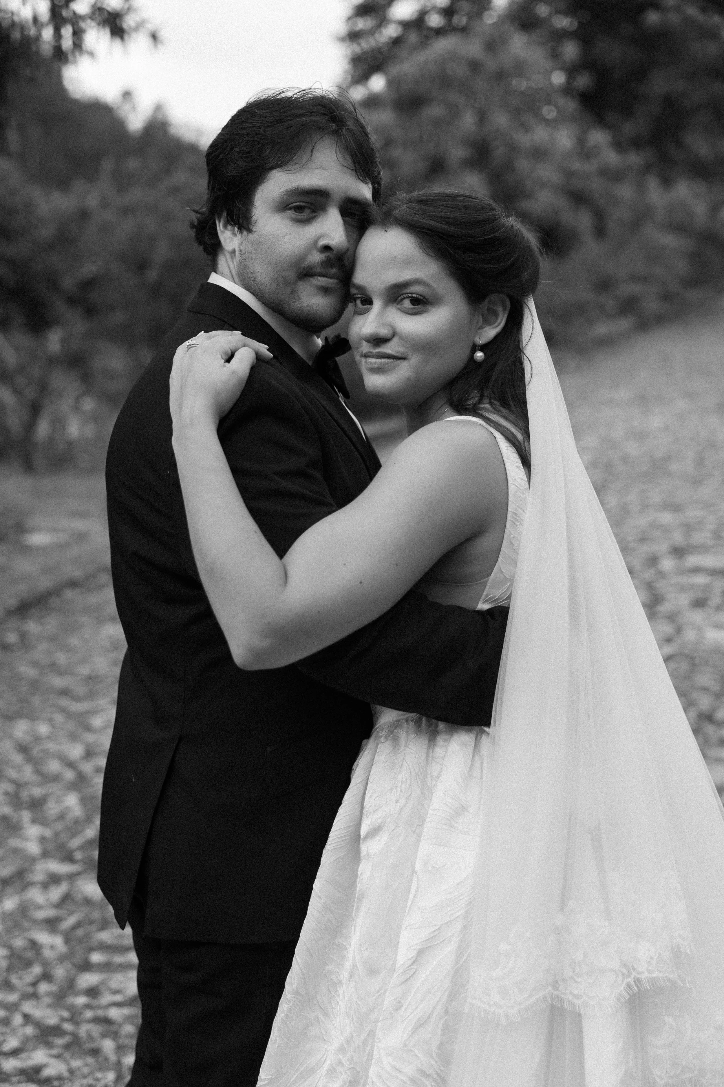 A black and white photograph of a bride and groom hugging outdoors, with trees in the background. The groom is wearing a tuxedo and the bride is in a wedding dress with a veil, both looking at the camera.