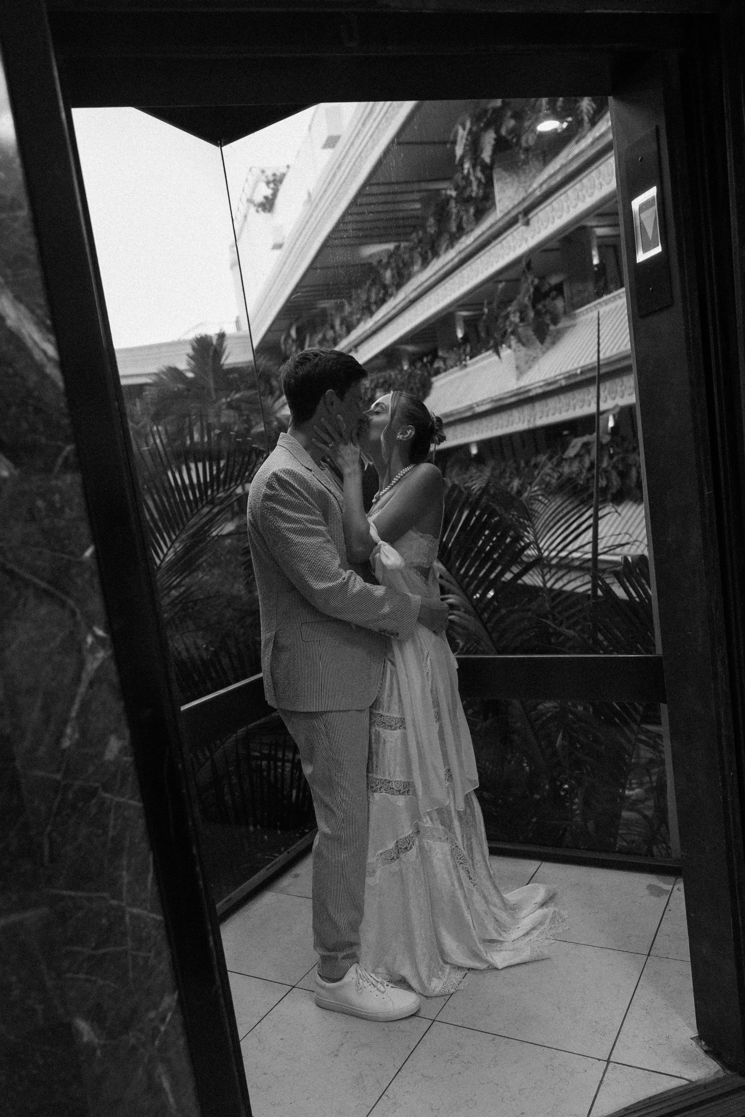 A black and white photo of a couple sharing a kiss, reflected in a mirror. The man is wearing a striped suit and sneakers, and the woman is in a long dress with lace details, accessorized with a pearl necklace. The reflection shows a building with mu