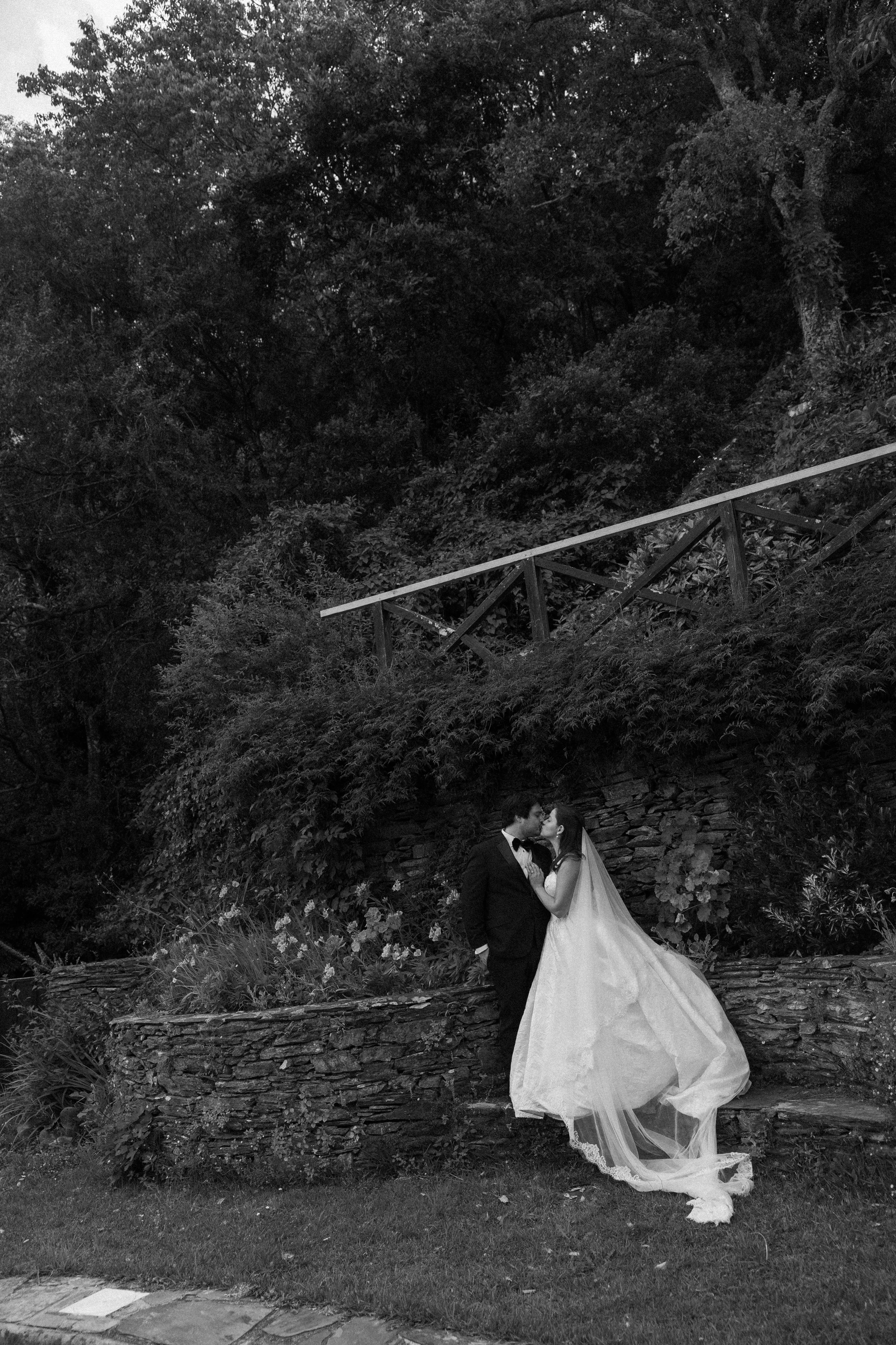 A bride and groom standing close together outdoors, surrounded by dense foliage and trees, with a wooden railing and stone wall in the background.
