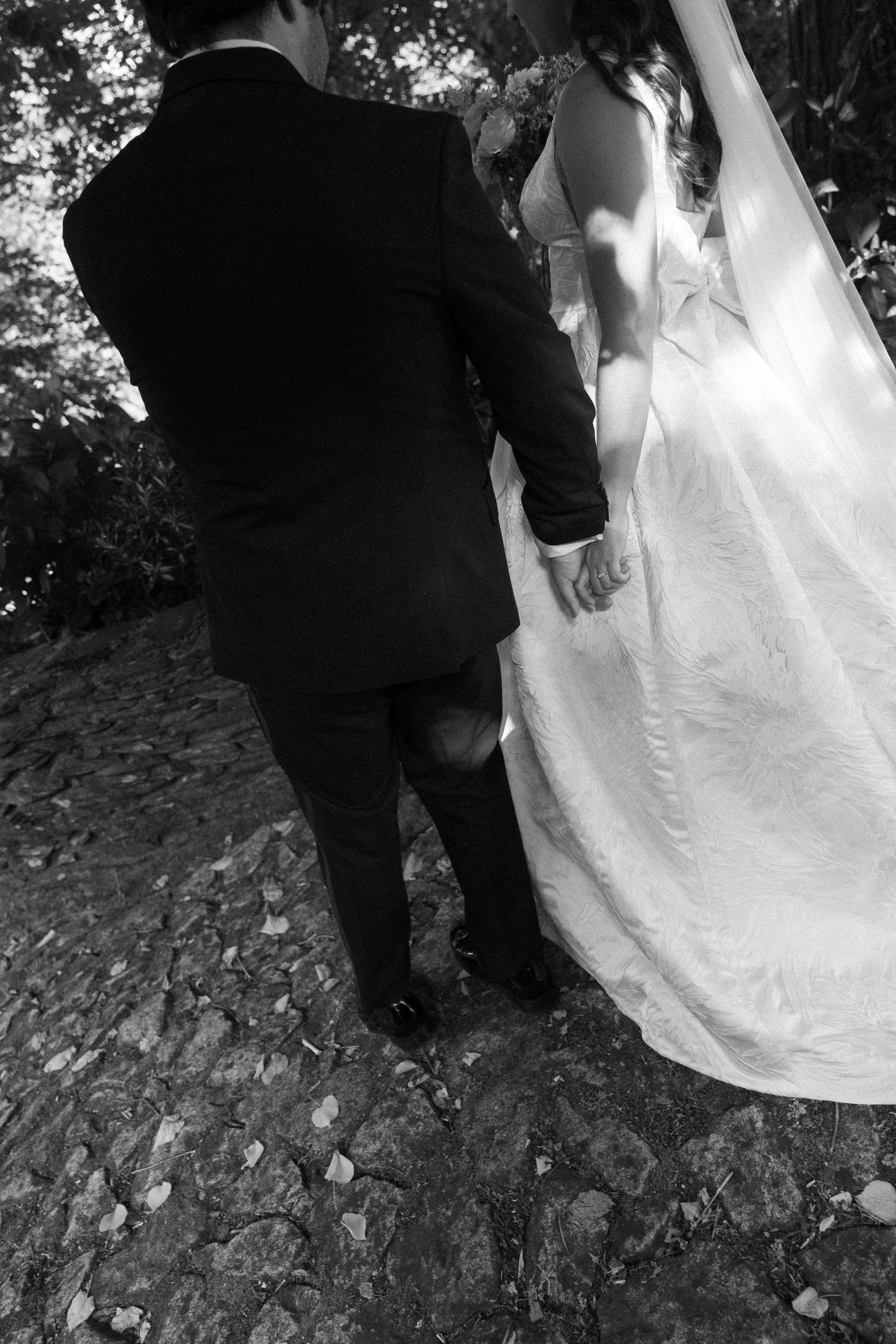 A black and white photo of a bride and groom holding hands during their wedding ceremony outdoors. The groom wears a suit, and the bride wears a wedding dress with a veil. The image is taken from behind and unposed.
