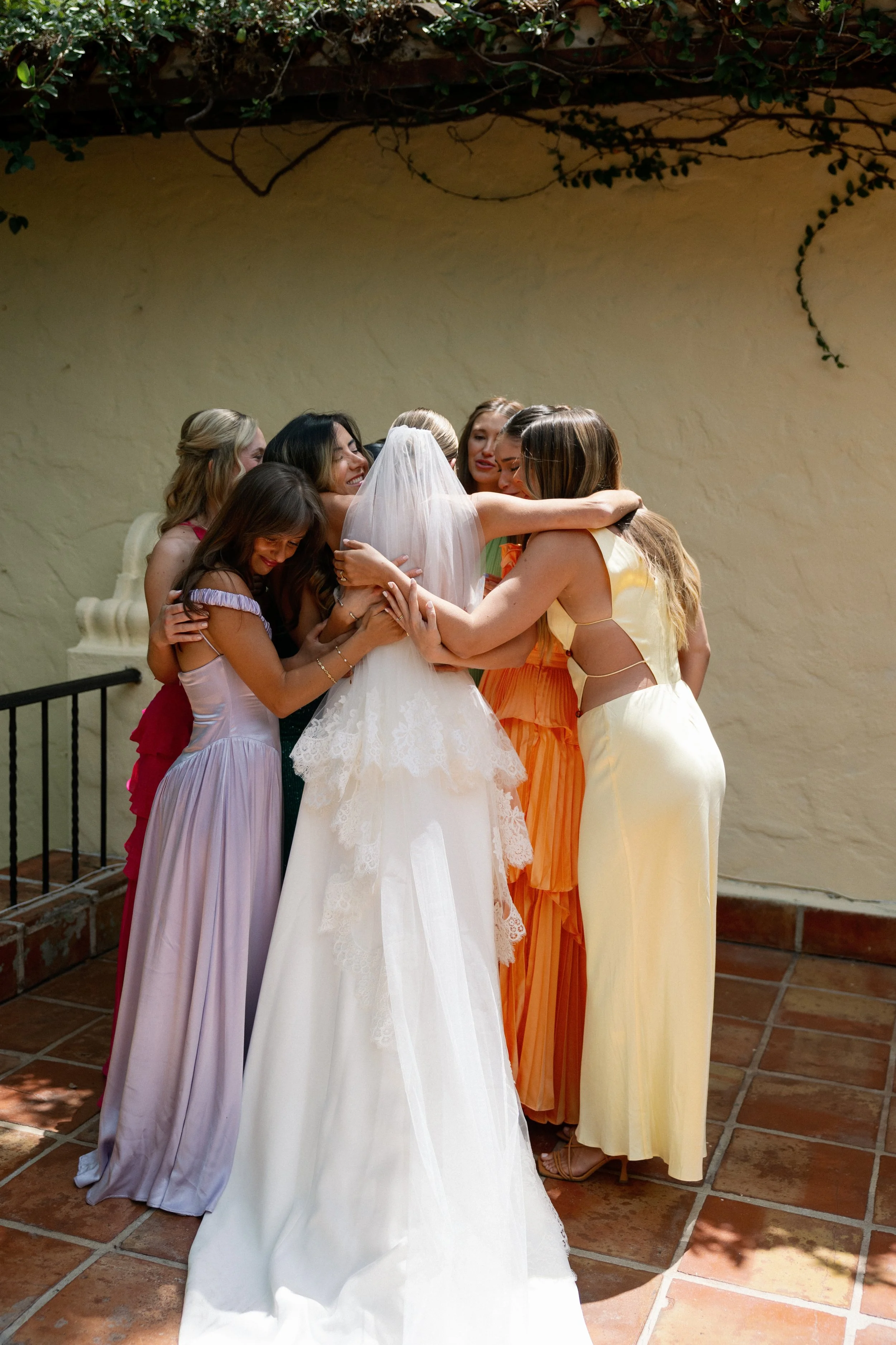 A group of women hugging at a wedding celebration, with the bride in a white wedding dress and veil, and the others in colorful dresses, on a tiled outdoor patio.