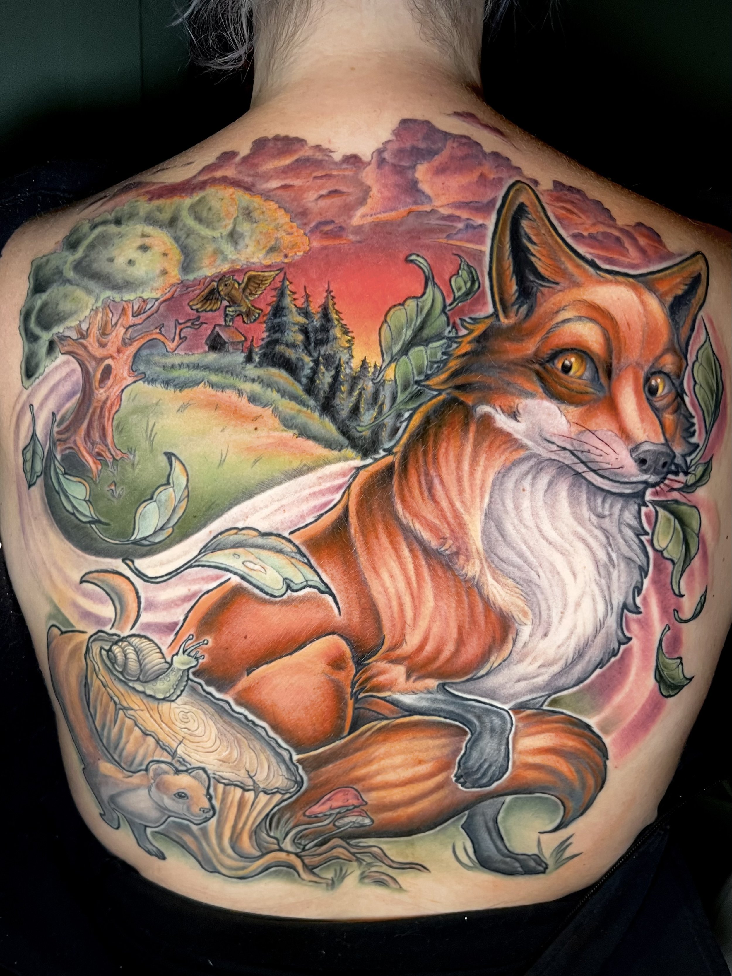 Colorful tattoo covering the person's back, featuring a fox in the foreground with detailed fur and expressive eyes, surrounded by leaves, trees, clouds, and stars, with a sunset or sunrise sky in the background.