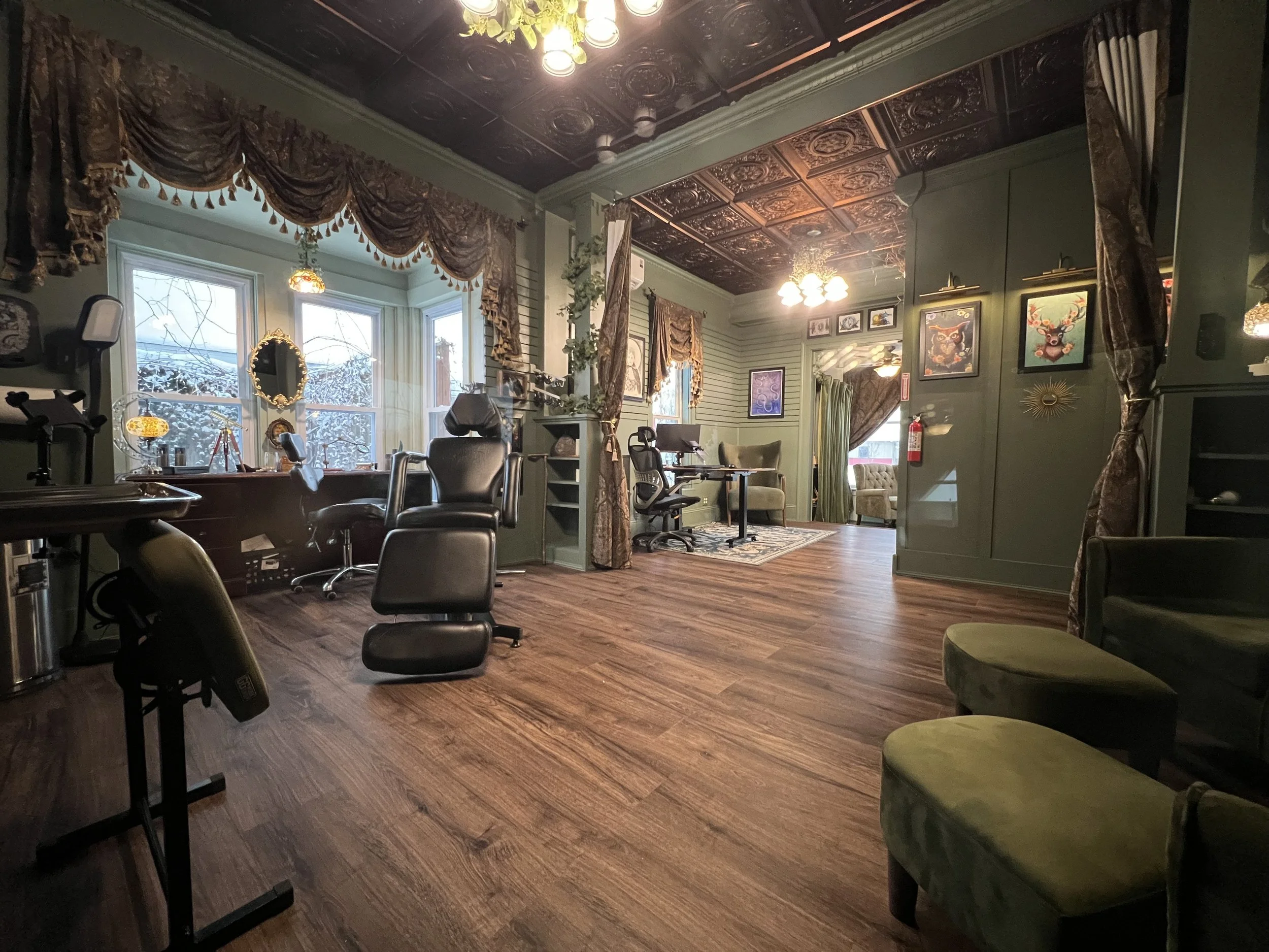 Interior of a cozy room with a green color scheme, featuring large windows with ornate curtains, multiple chairs, artwork on the walls, and warm lighting, with a view of snow outside.