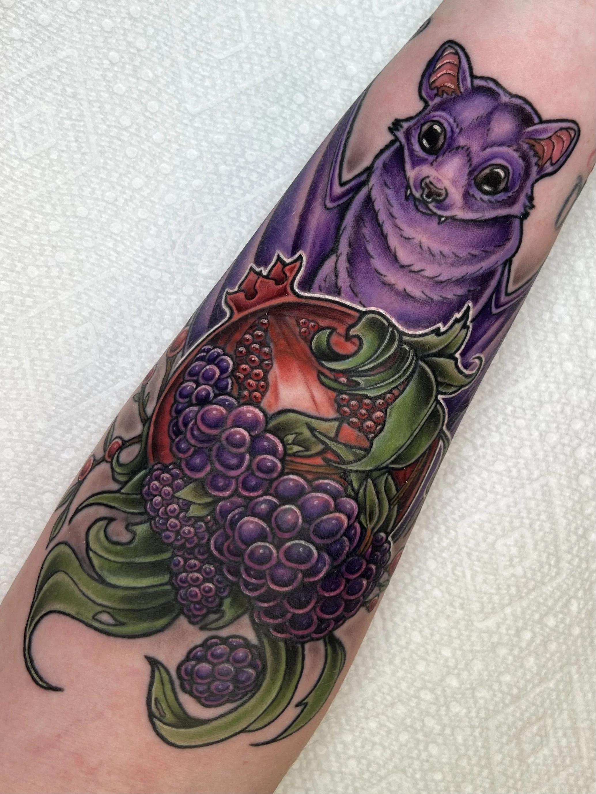 Colorful tattoo of a purple bat with black eyes, surrounded by grape clusters and green leaves.