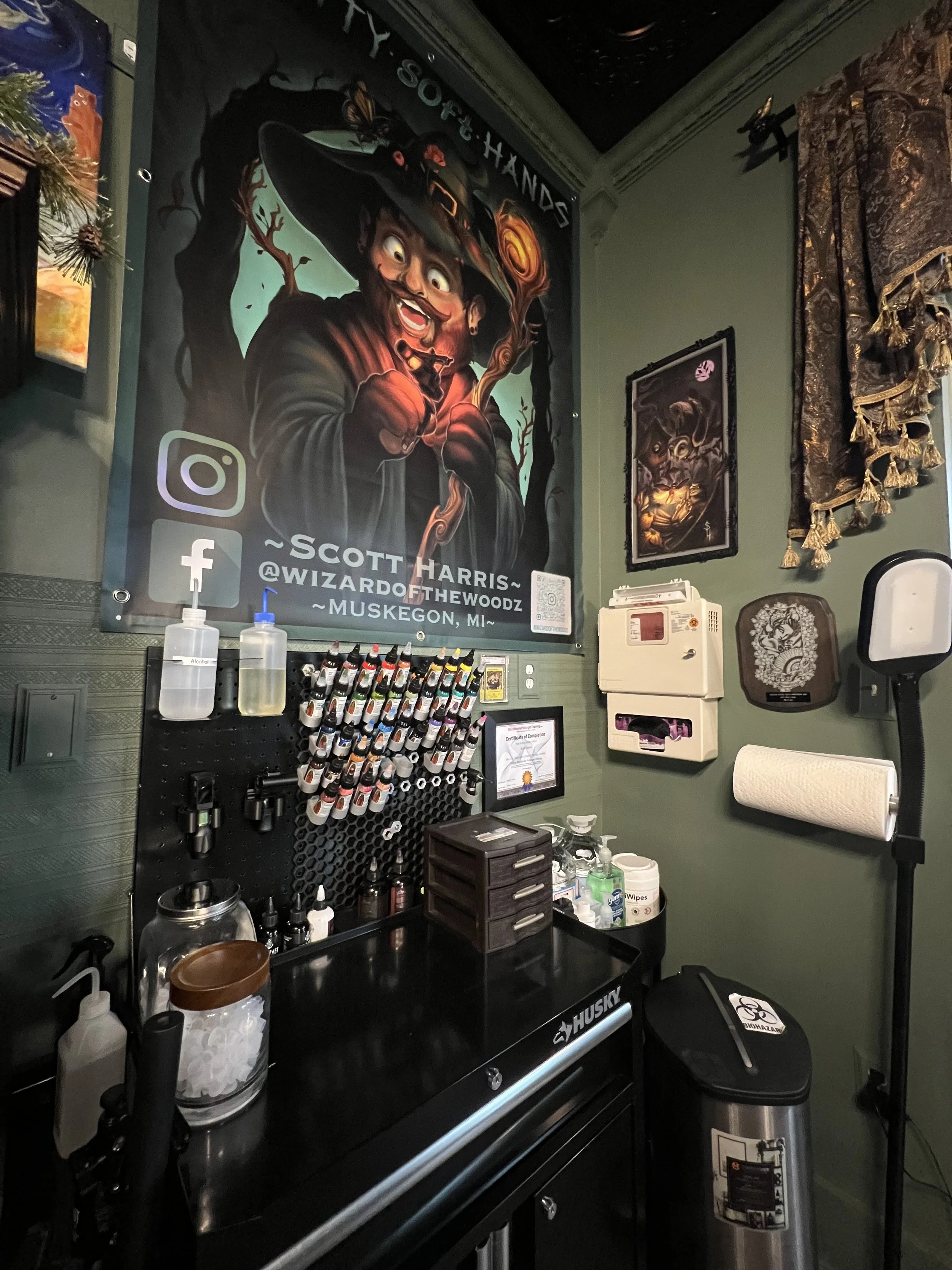 A tattoo shop workstation with tattoo ink bottles, disinfectant, hand sanitizer, and cleaning supplies on a black counter. There is a tattoo machine on a pegboard, framed art, and a large colorful poster of a witch with a hat and a staff on the wall. The poster includes social media handles and location details.