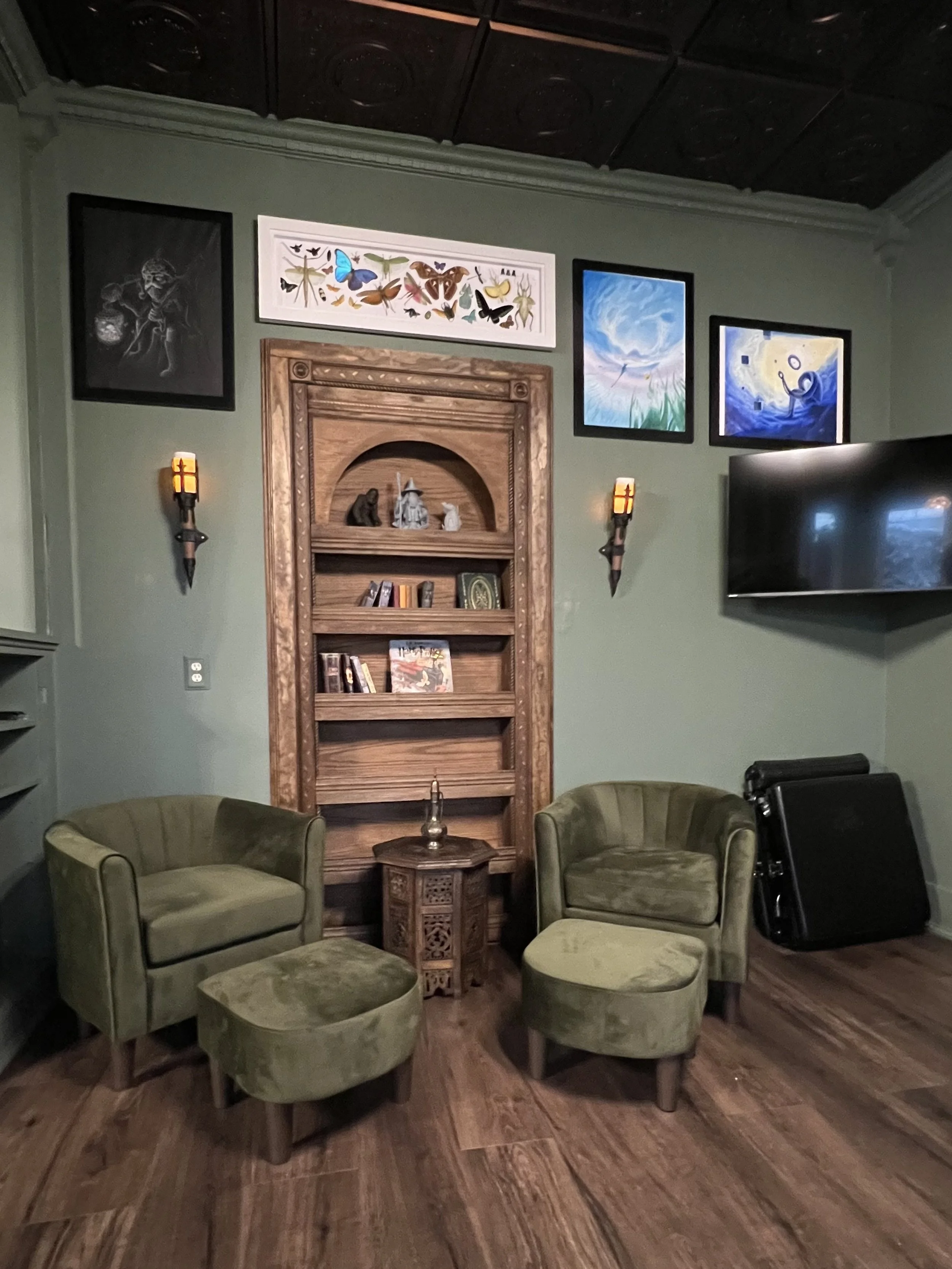 A room with two green armchairs and a matching ottoman arranged in a semi-circle around a small wooden table. A built-in wooden bookshelf with decorative items and books is on the wall behind them. The wall has framed artwork including a butterfly illustration, a blue sky with grass, and a blue abstract piece. There are also two wall sconces with warm lighting on either side of the bookshelf. A large flat-screen TV is mounted on the wall to the right of the artwork. The room has dark wood flooring and a dark wooden ceiling.