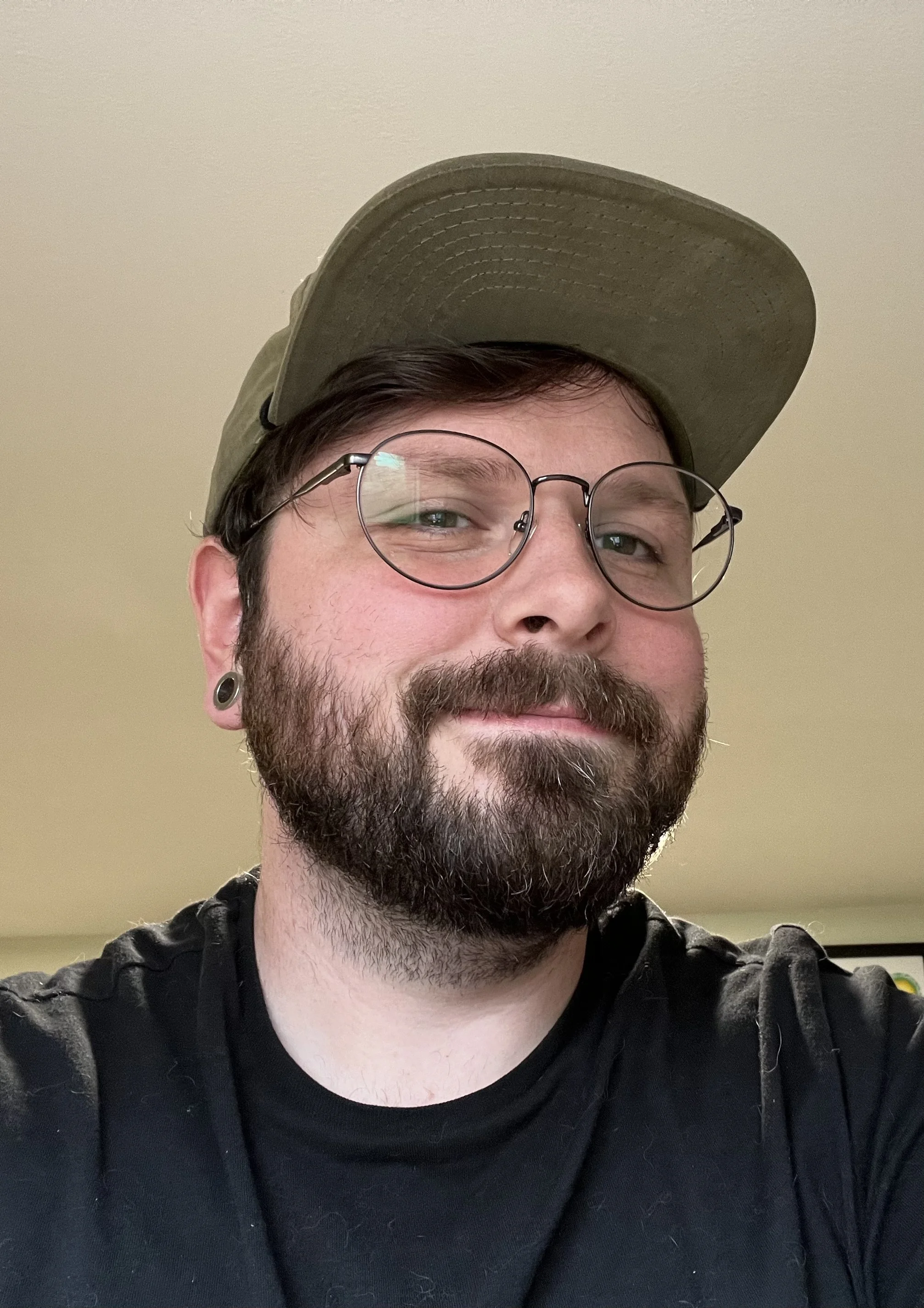A man with glasses, a beard, and a mustache wearing a green cap and a black shirt, smiling with a neutral background.
