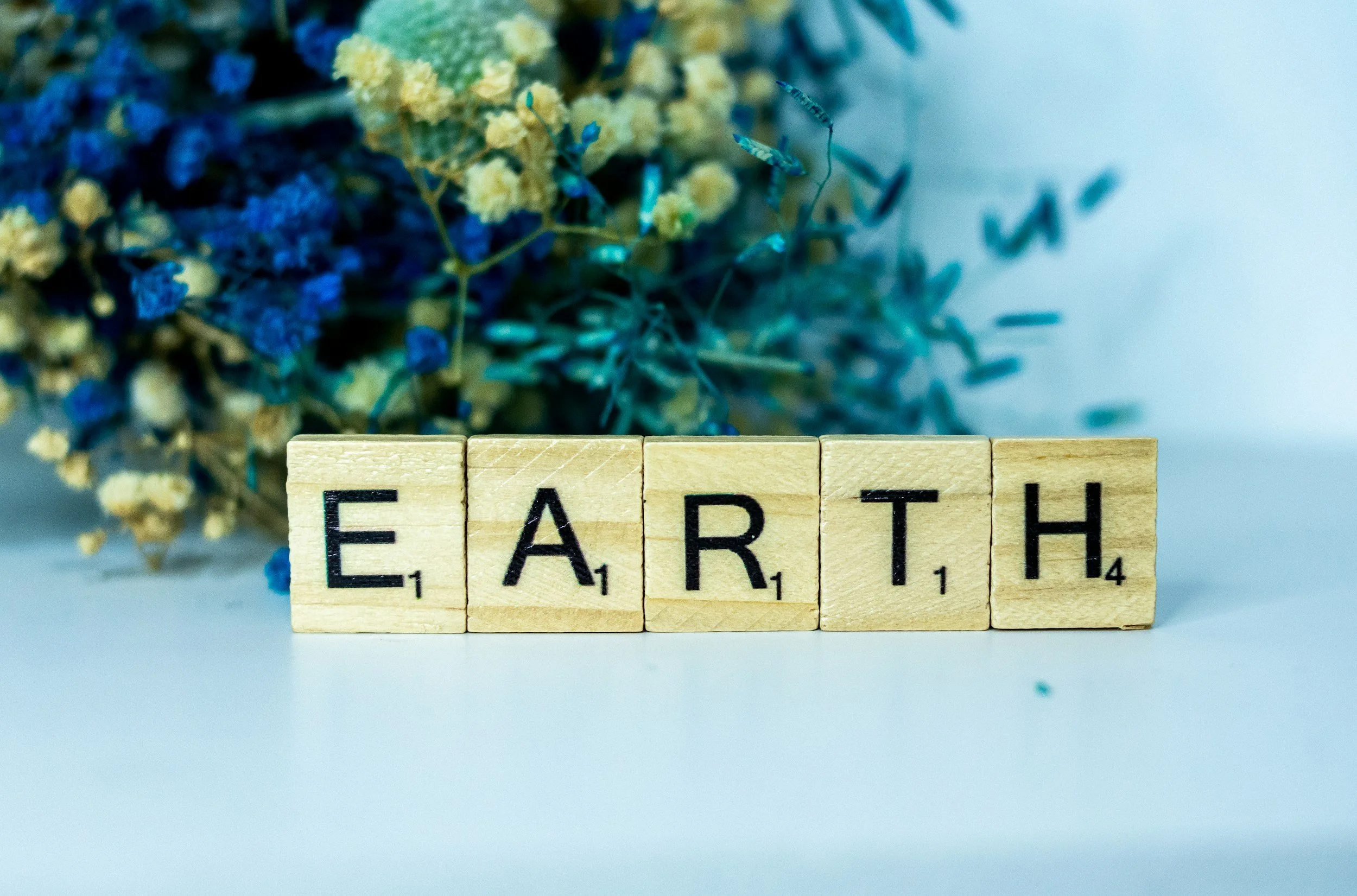 Wooden Scrabble tiles spelling out 'EARTH' with a bouquet of blue and yellow flowers in the background.