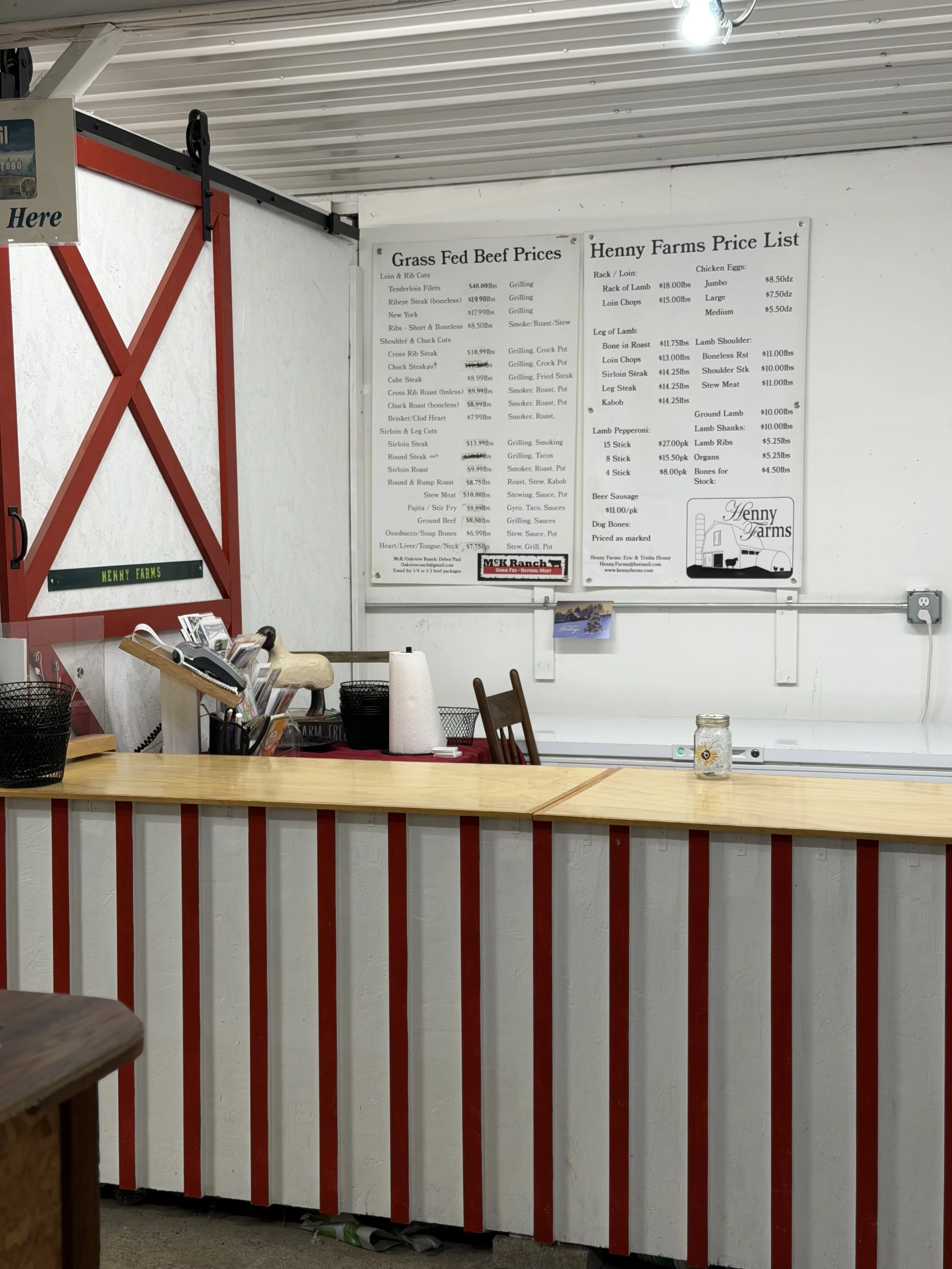 Interior of a farm shop or butcher shop with white walls and red decorative paneling, displaying two boards listing beef and lamb prices, along with some hanging lights and shelves with miscellaneous items.
