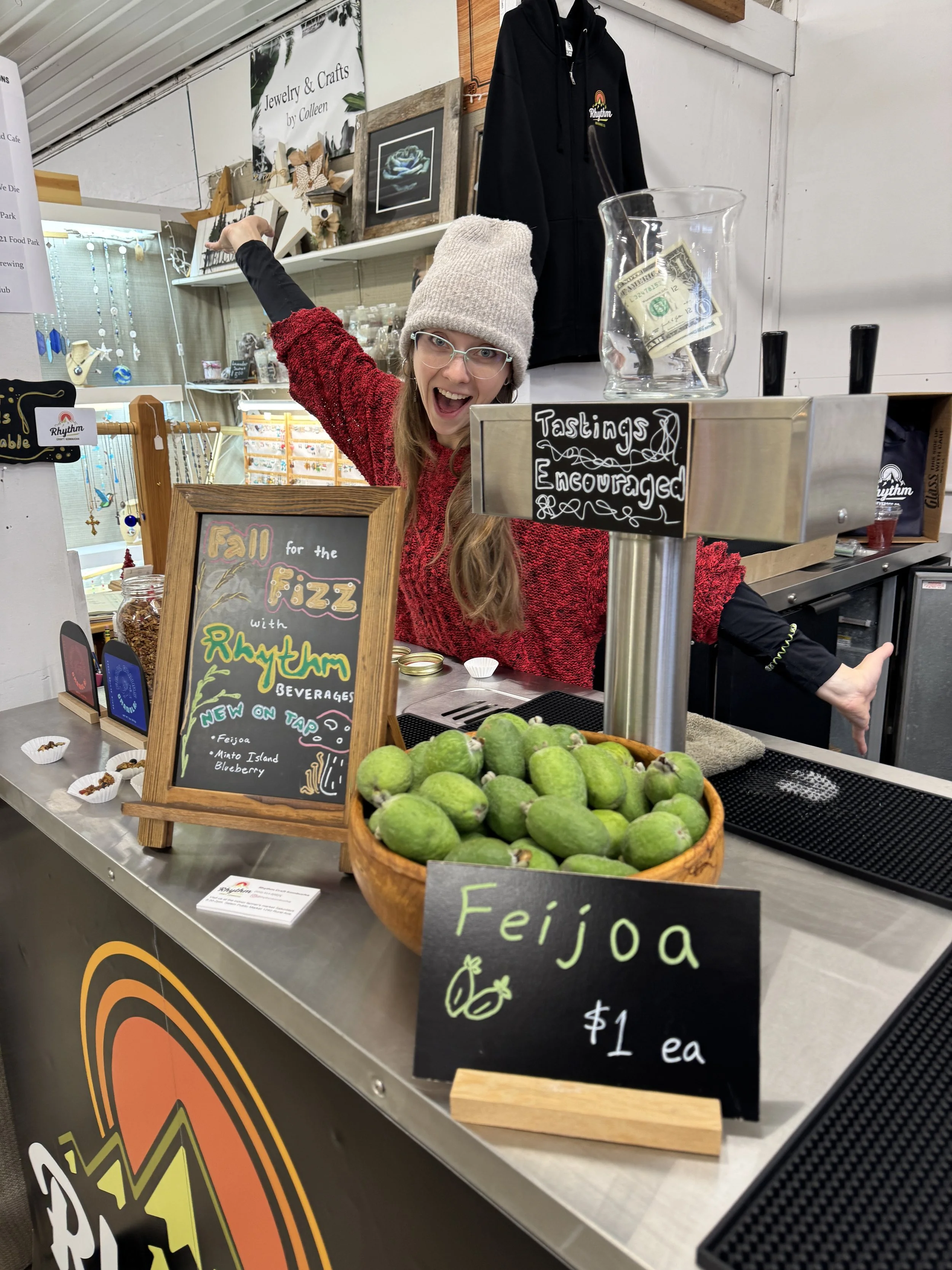 A woman wearing glasses, a beige beanie, and a red sweater is smiling and gesturing with her arm extended at a booth selling Feijoa fruits. The booth has a chalkboard sign advertising Feijoa for $1 each, and a basket of green Feijoa fruits in the foreground. There are various signs and decorations, including a black hoodie hanging on the wall and a glass jar with dollar bills.