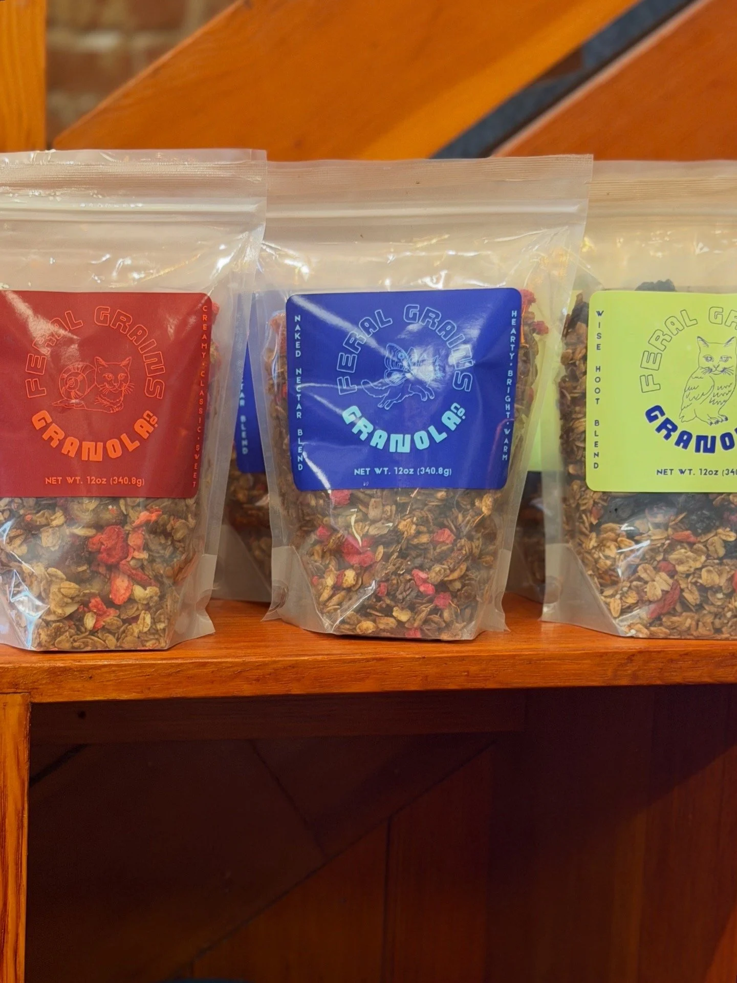 Three bags of trail mix on a wooden shelf, each with different colored labels: red, blue, and yellow.