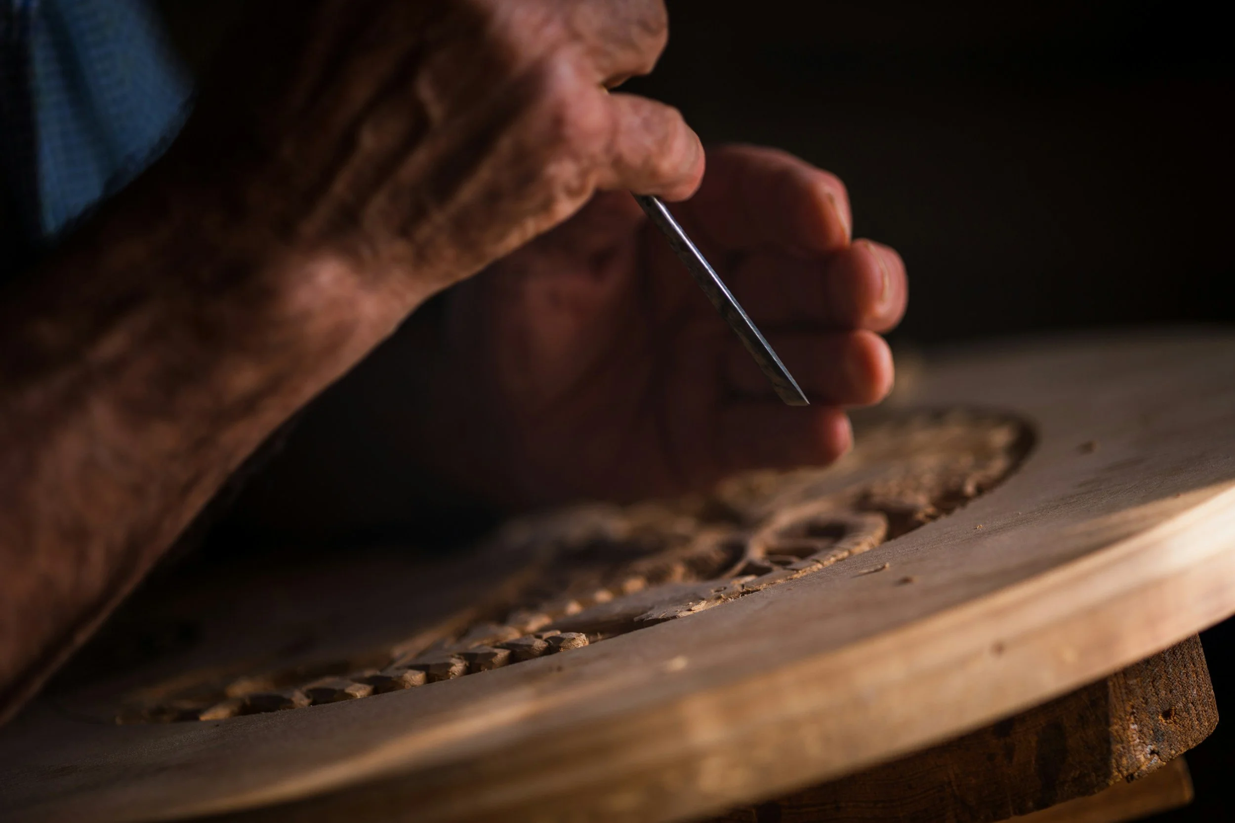A close-up of hands using a small carving tool on a piece of wood, with wood shavings around.
