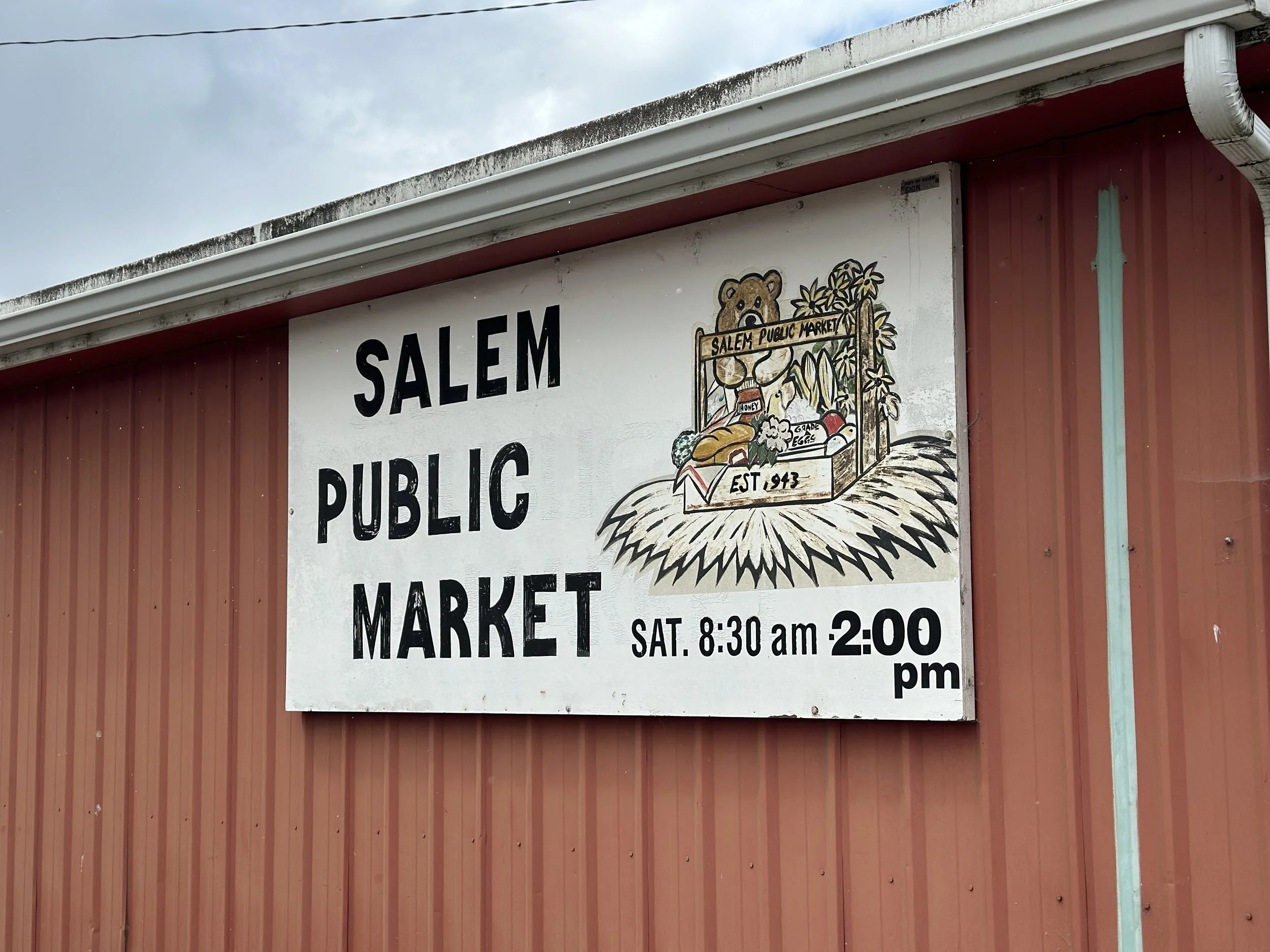 A sign on a red building that reads 'Salem Public Market' with additional details about the market hours. There is a painted image of a bear's face and a basket of produce with the words 'Salem Public Market' and 'Est. 1943'.