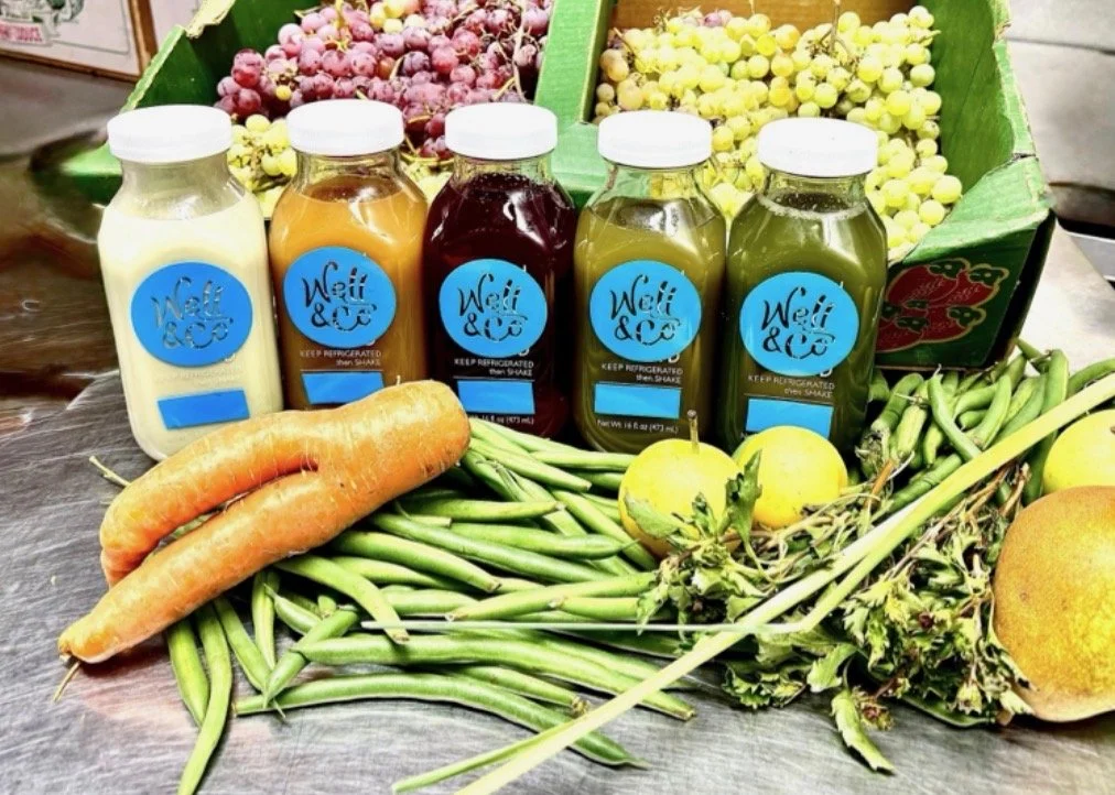 A display of five bottled juices labeled 'Welli & Co' surrounded by fresh vegetables and fruits, including carrots, green beans, apples, and grapefruit, with boxes of grapes in the background.