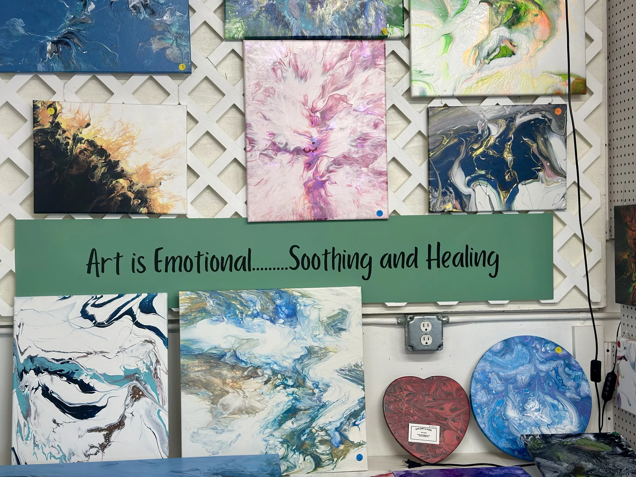 Display of abstract paintings in various colors and styles arranged on a white lattice wall with a sign that reads 'Art is Emotional... Soothing and Healing'.