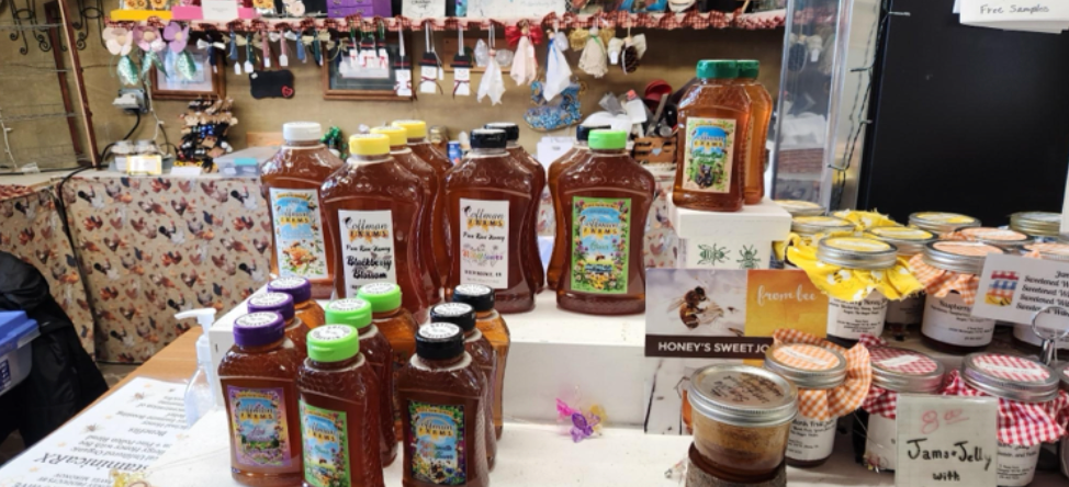 Display of honey bottles and jars at a market stall, with various labels and decorative items in the background.