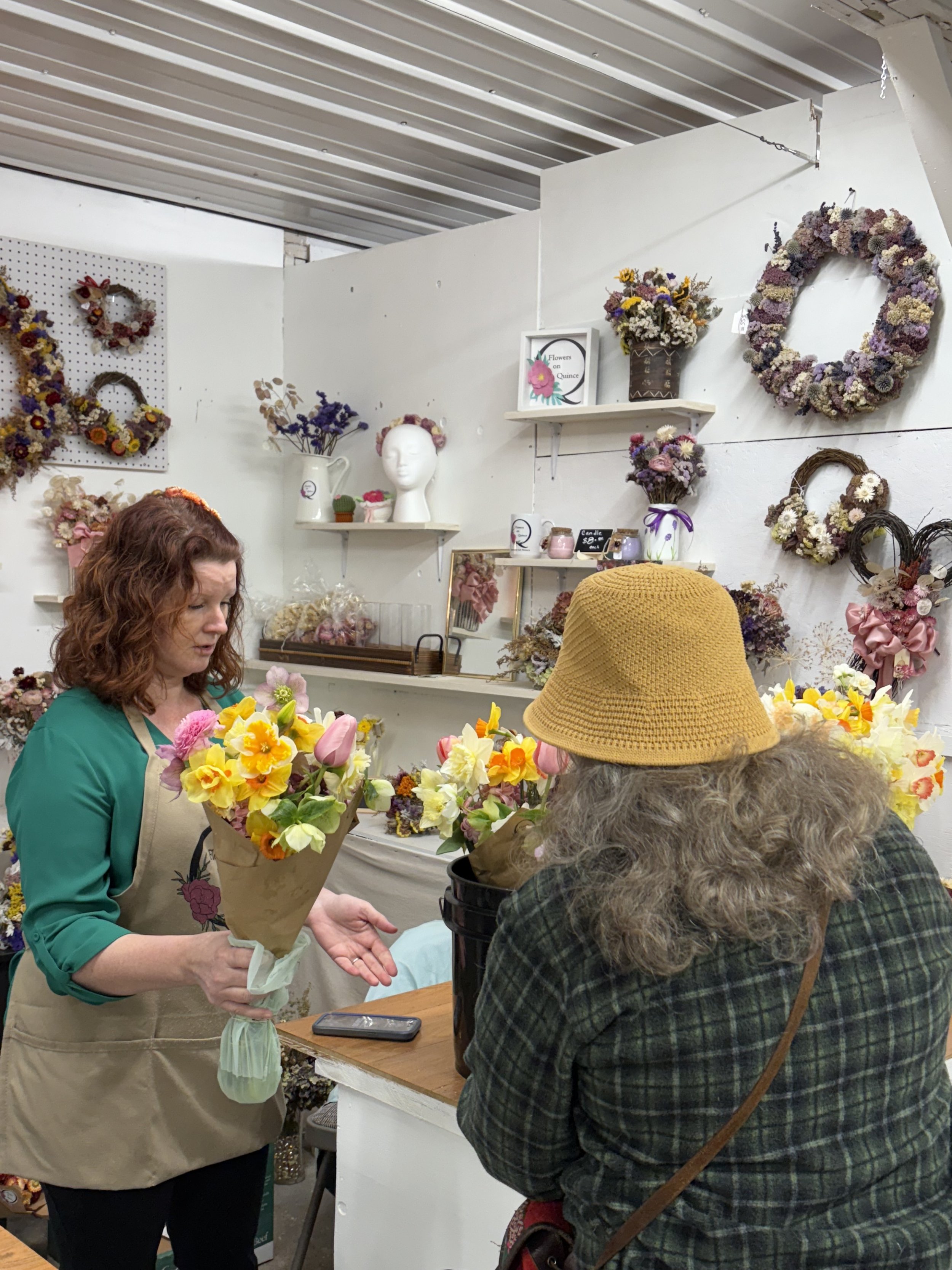 A woman with curly red hair wearing a green top and beige apron handing a bouquet of yellow and pink flowers to an older woman wearing a checkered jacket and a mustard yellow hat. They are inside a shop with white walls decorated with flower wreaths and arrangements.