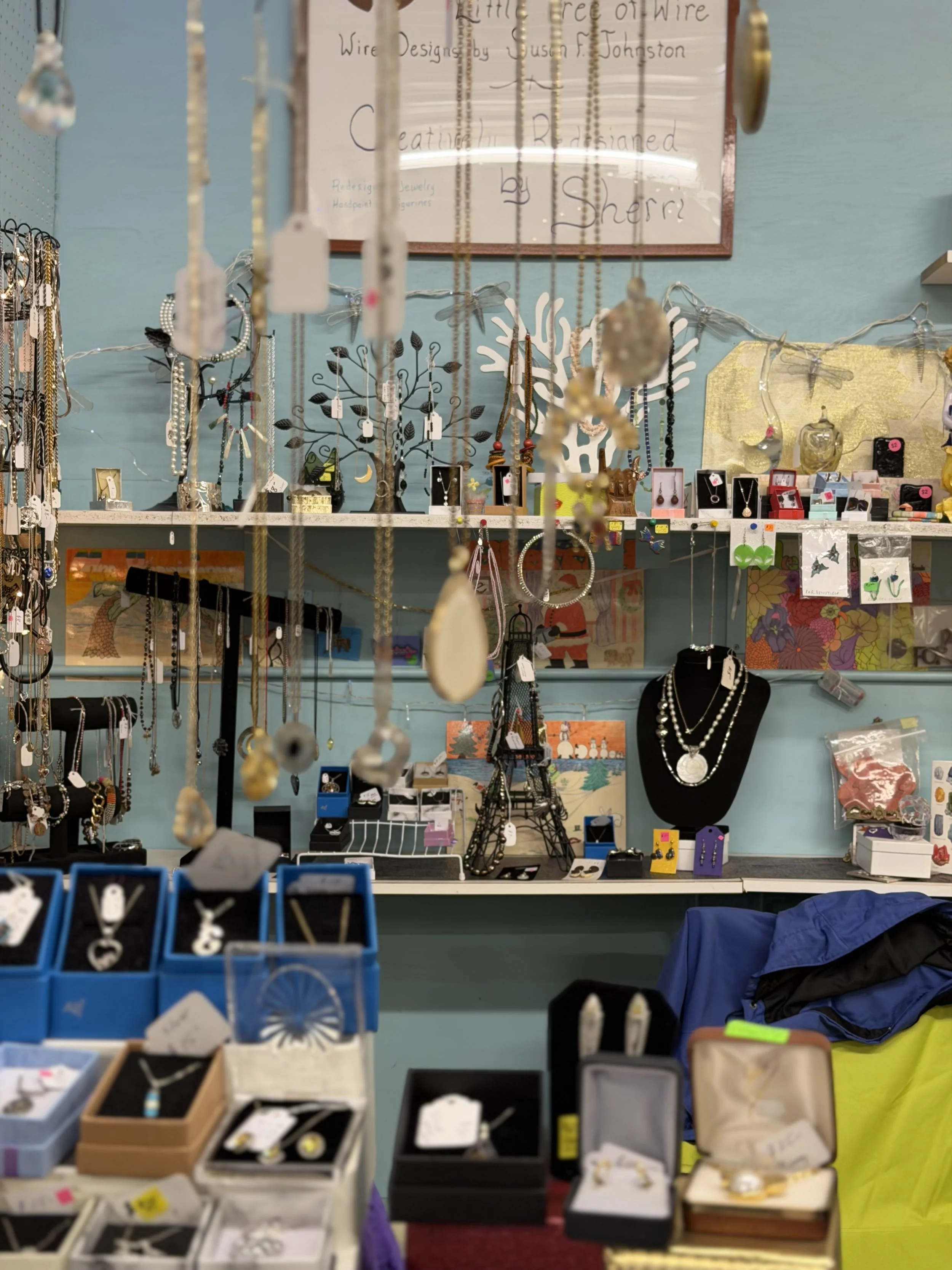 A jewelry display table with various necklaces, earrings, and rings arranged in boxes and on a necklace bust, with a backdrop of shelves and a framed sign in the background.