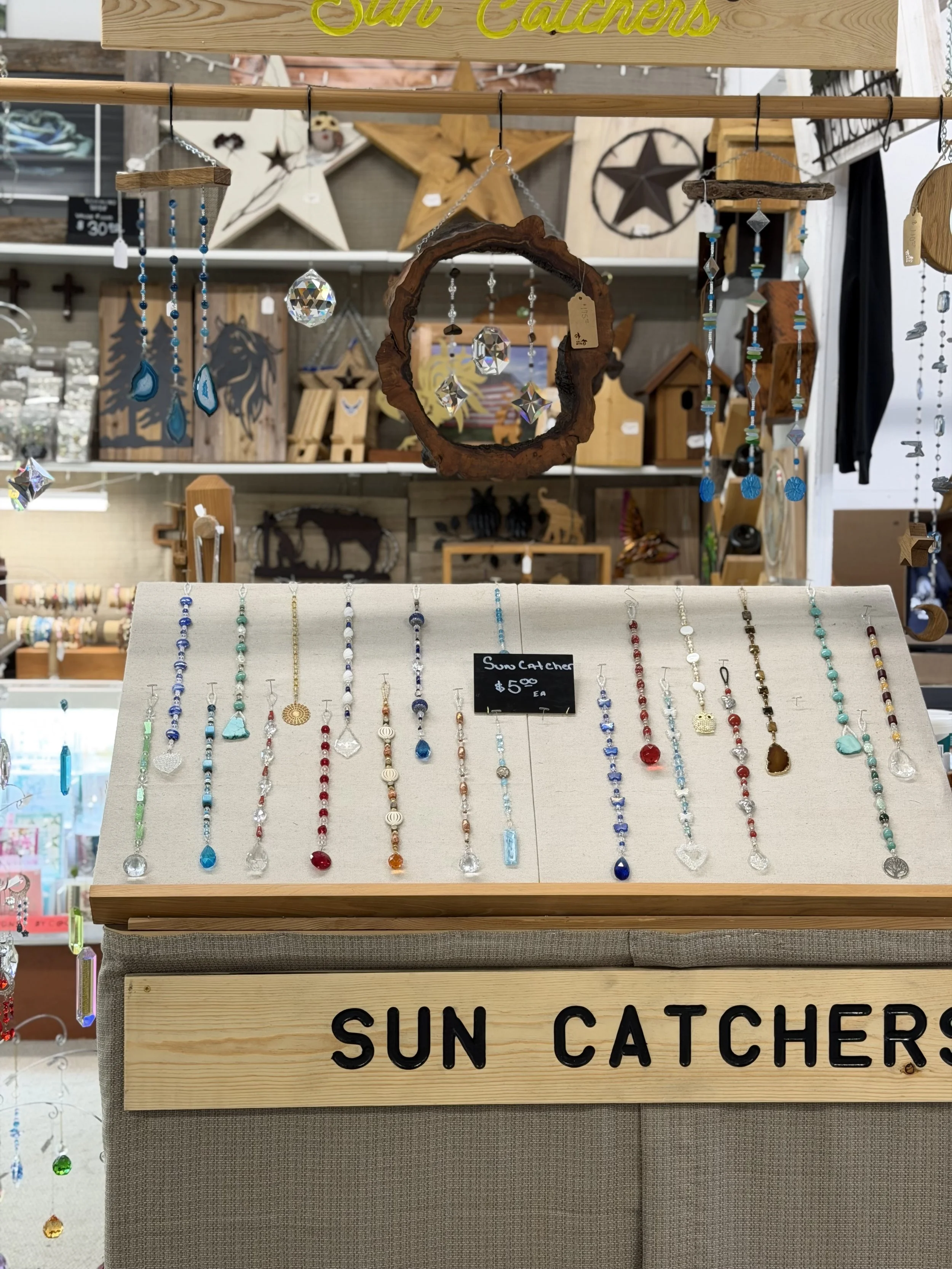 Display of beaded necklaces hanging on a white board with a sign that reads 'Sun Catcher' and priced at $5.00 each. The background shows decorative wooden stars and other wooden decor items.