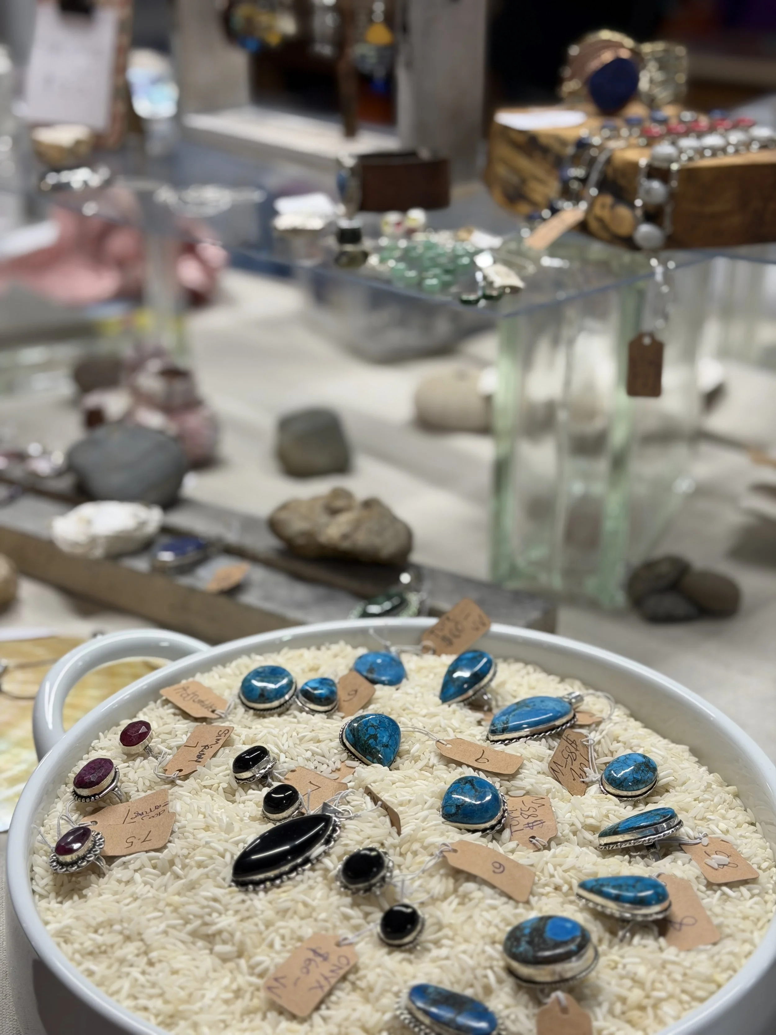 A display of various silver and turquoise jewelry, including rings and pendants, on a bed of white rice, with handwritten price tags attached. In the background, there are rocks and jewelry boxes.