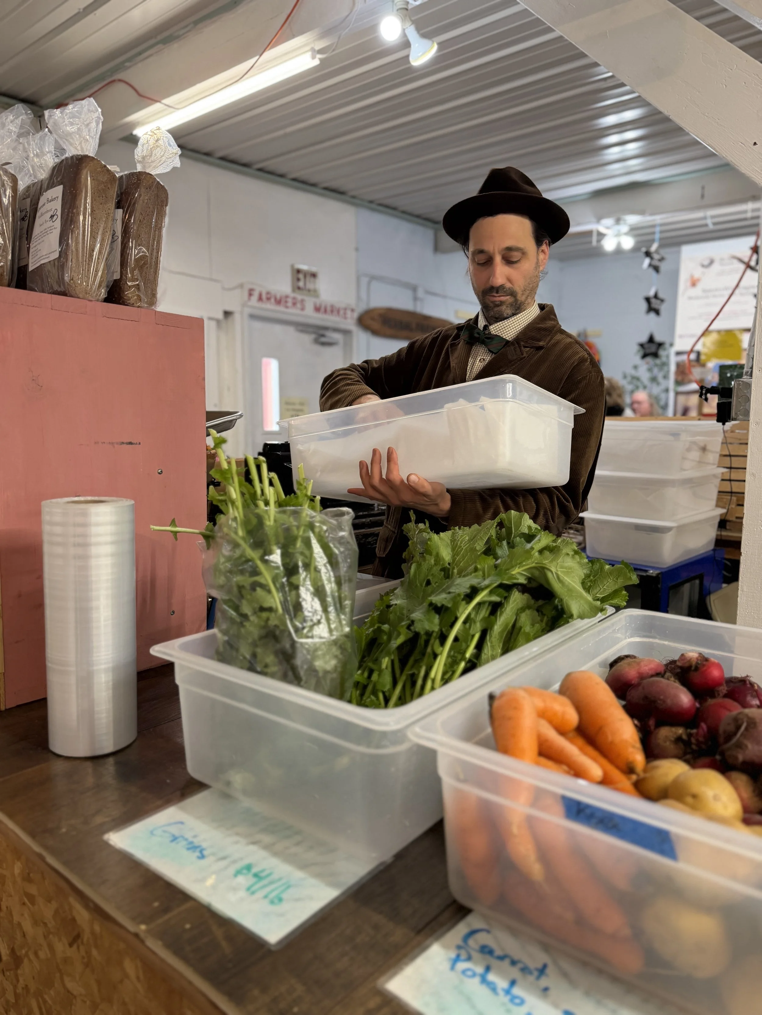 A man wearing a brown jacket, patterned shirt, green bowtie, and black hat shopping at a produce stand. He is holding a plastic container, and there are fresh vegetables such as carrots, radishes, and potatoes in clear plastic bins on the counter. The setting appears to be indoors, possibly at a farmers market.