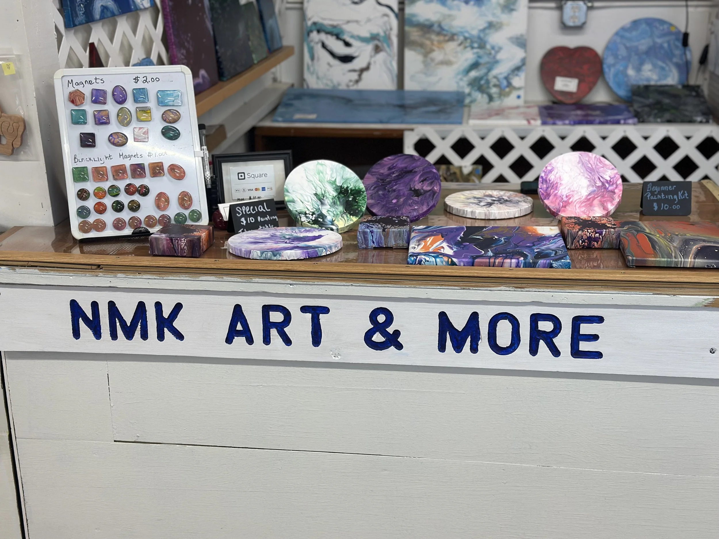 Painted circular wooden art pieces with colorful abstract designs displayed on a table at NMK Art & More. A white sign on the table indicates prices for magnets and painting kits.