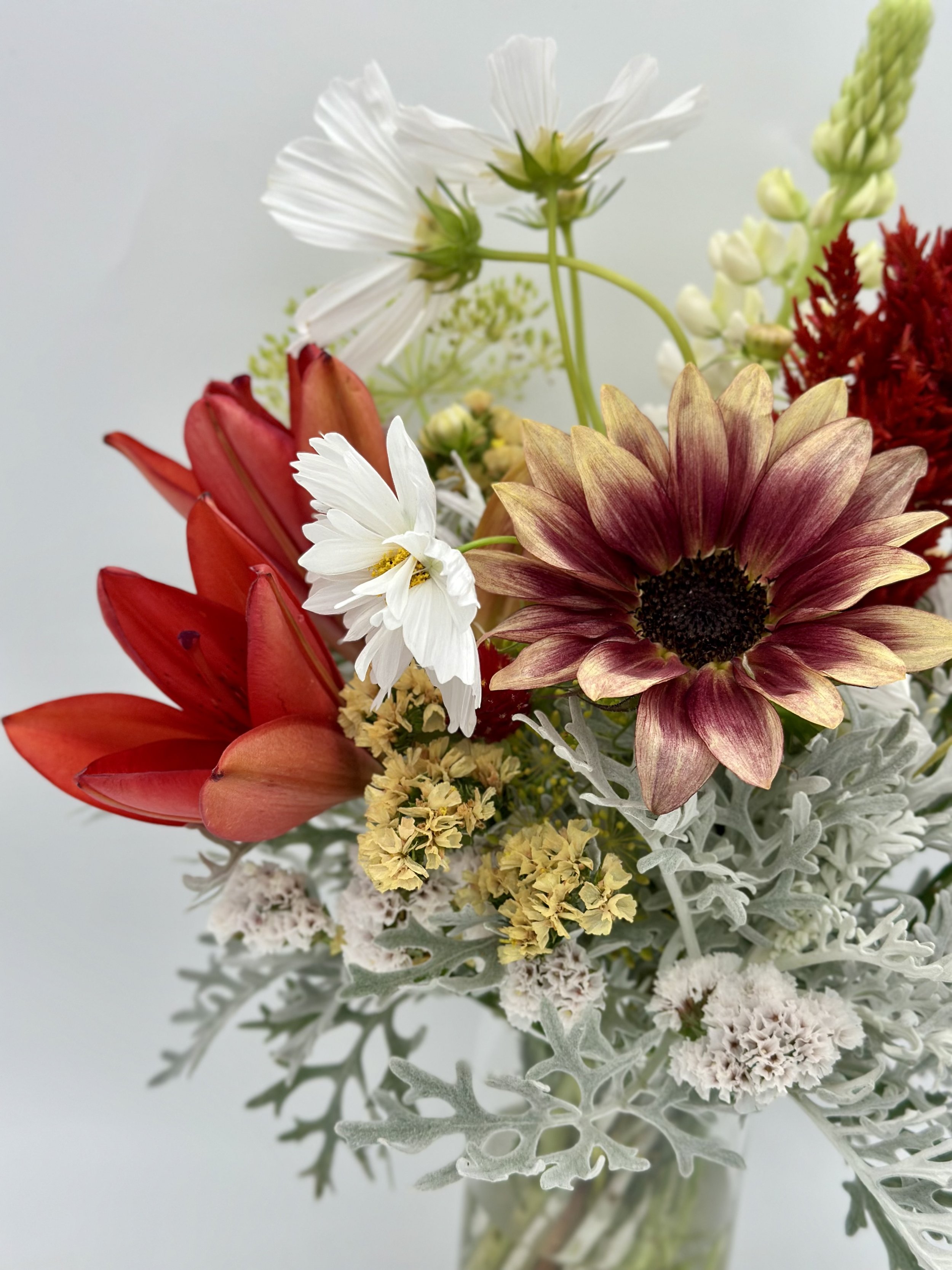 A close-up view of a floral arrangement featuring various flowers including white daisies, red lilies, a large Burgundy and yellow sunflower, and other smaller flowers, set against a light background.