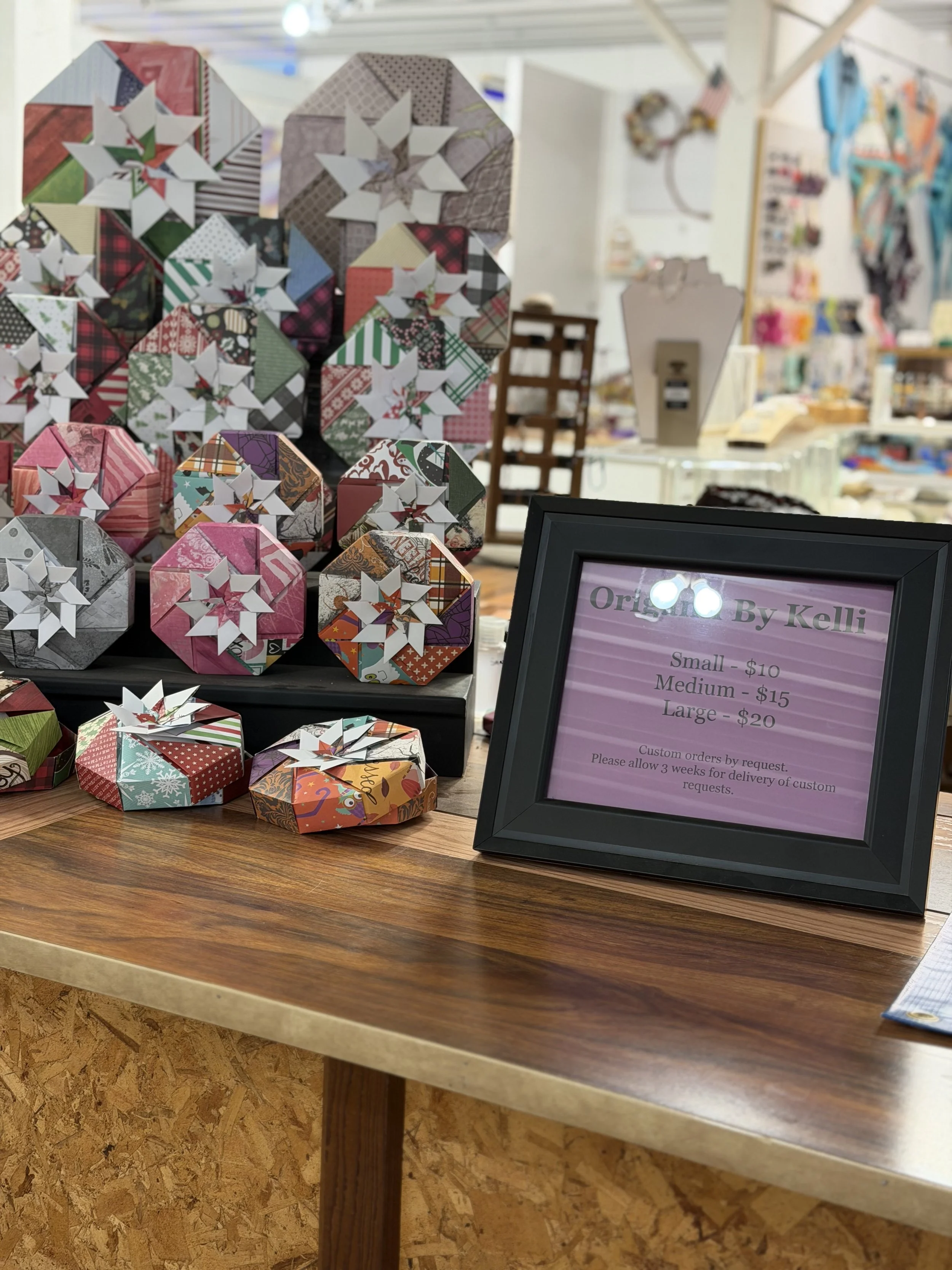 Display of origami paper stars decorated with Christmas-themed paper, arranged on a table at an indoor market or craft fair. A framed sign lists prices for small, medium, and large origami stars and mentions custom orders.