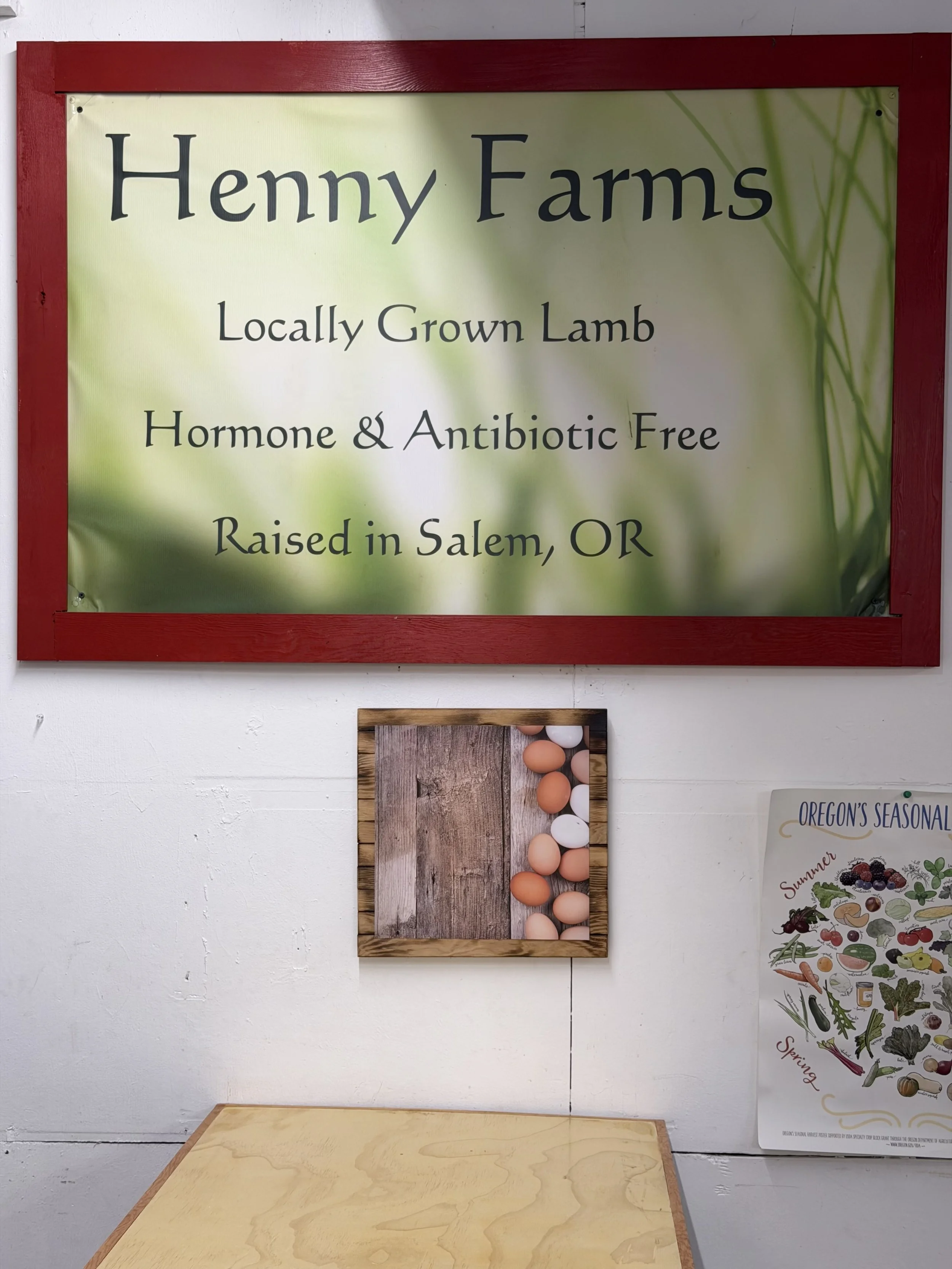 A sign reading "Henny Farms, Locally Grown Lamb, Hormone & Antibiotic Free, Raised in Salem, OR" on a red-framed display board. The background is a green and light-colored design. Below the sign is a wooden table and partially seen poster with various vegetables.