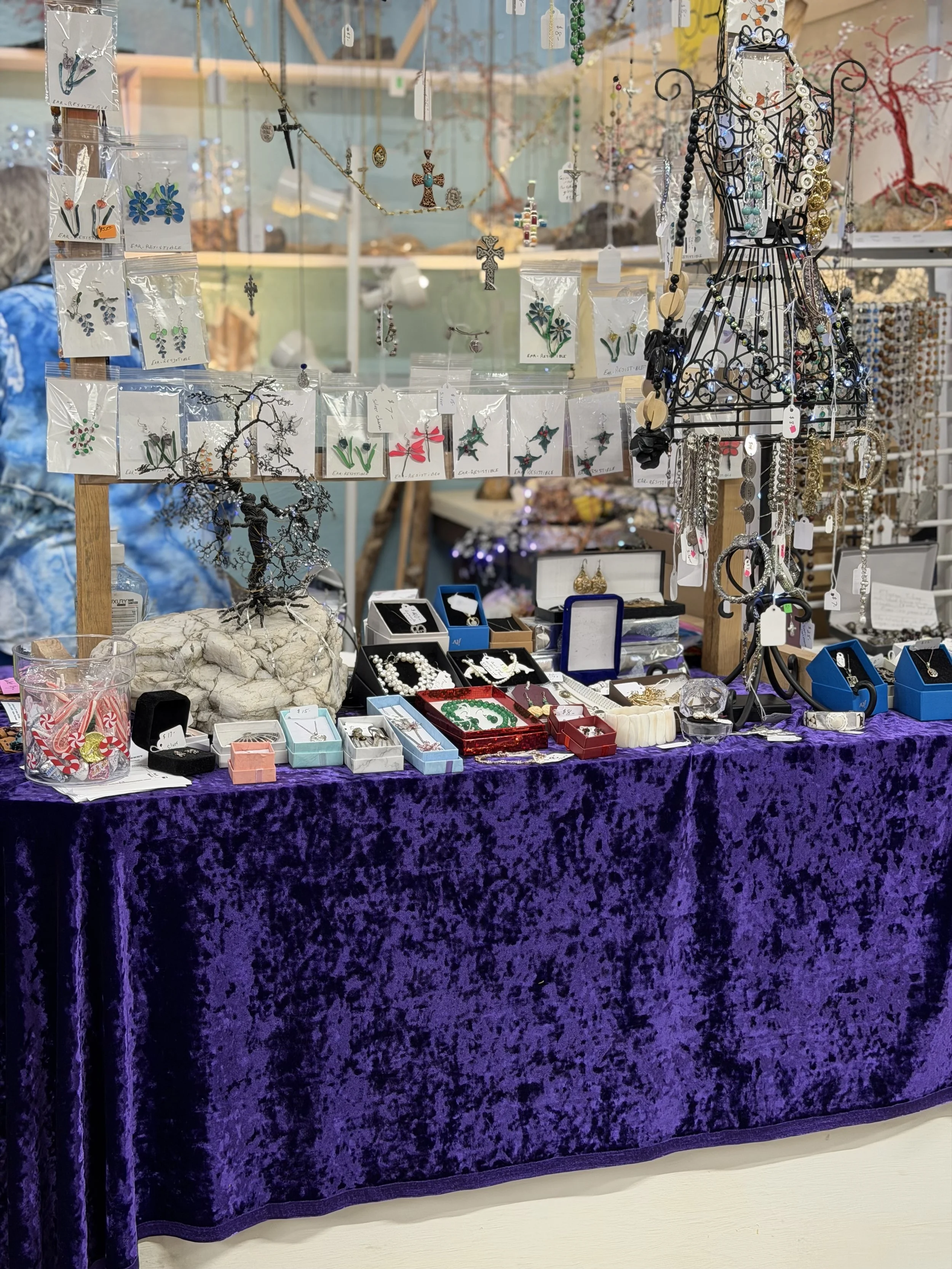 Display table covered with purple velvet cloth showcasing jewelry items, including necklaces, earrings, and rings. Jewelry is arranged in small boxes, on stands, and hanging from a black wire dress form. The background features a glass partition with additional jewelry and decorative items.