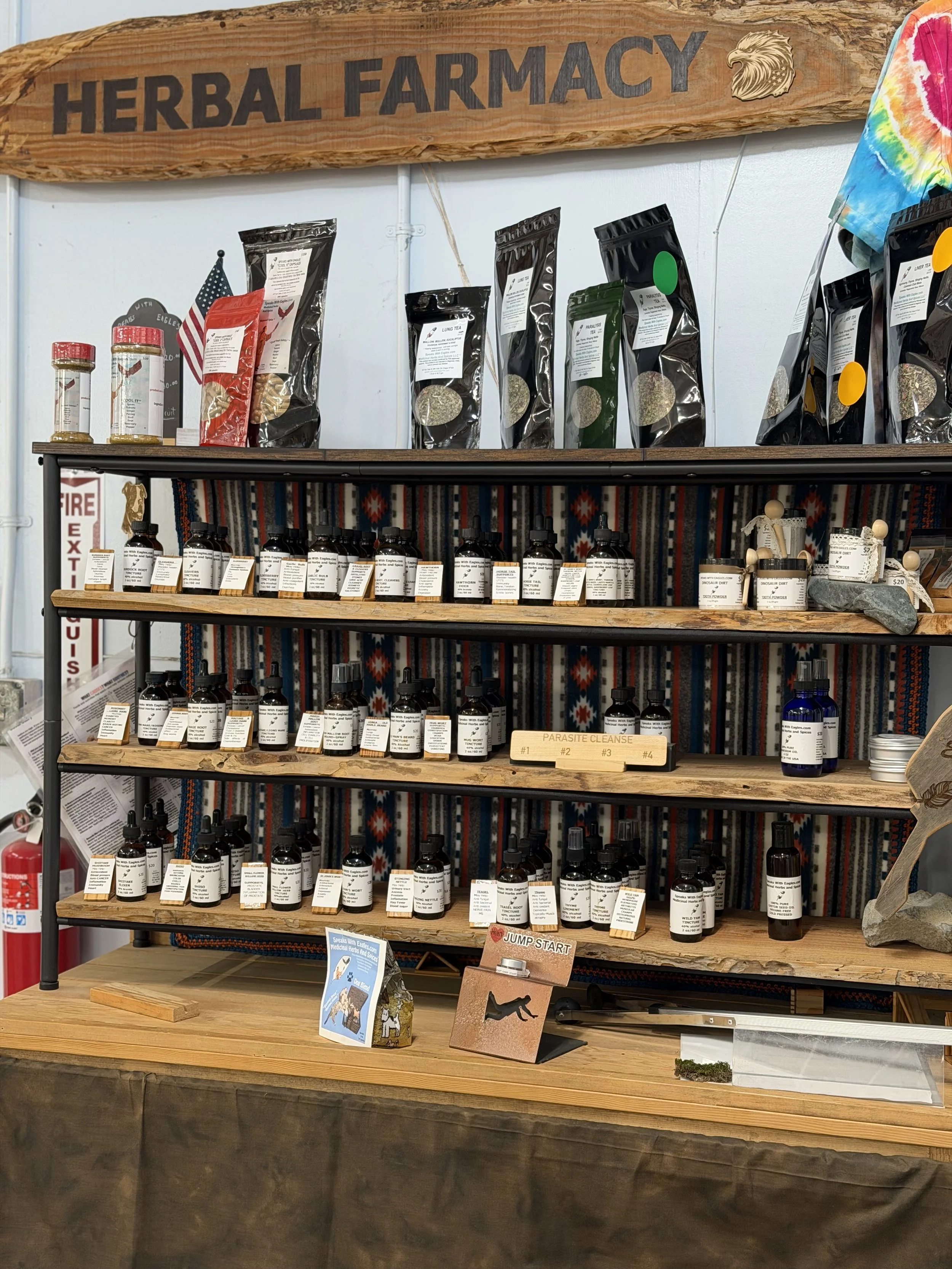 Display shelf with herbal products in black bottles, informational cards, and decorative items with a rustic wooden sign reading 'Herbal Farmacy' and an eagle logo. A small American flag and various labeled bottles are also visible.
