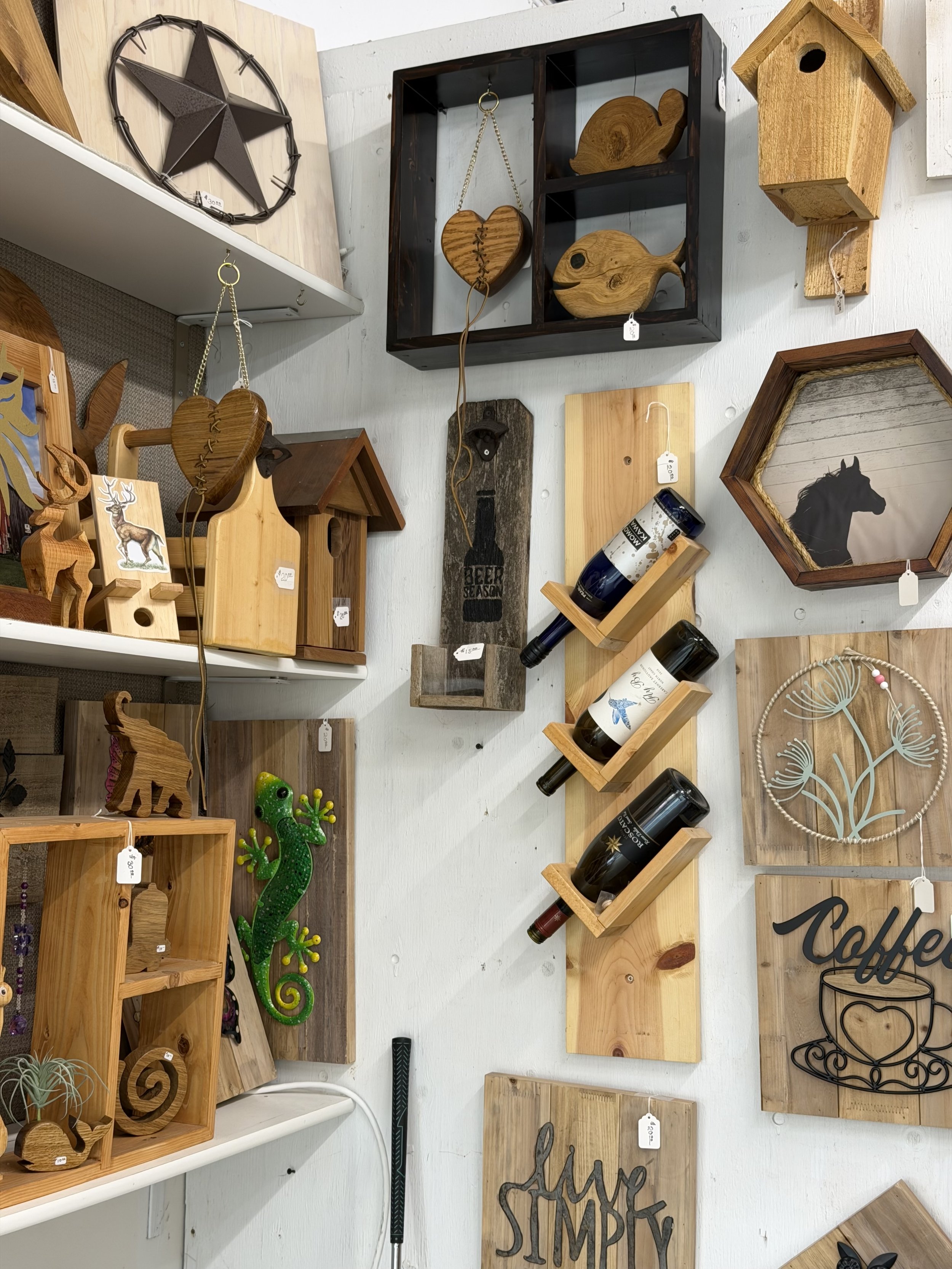 A display of various decorative wooden items and artwork, including a star wall hanging, fish-shaped decor, a gecko ornament, and wooden signs with text, arranged on white shelves and wall.
