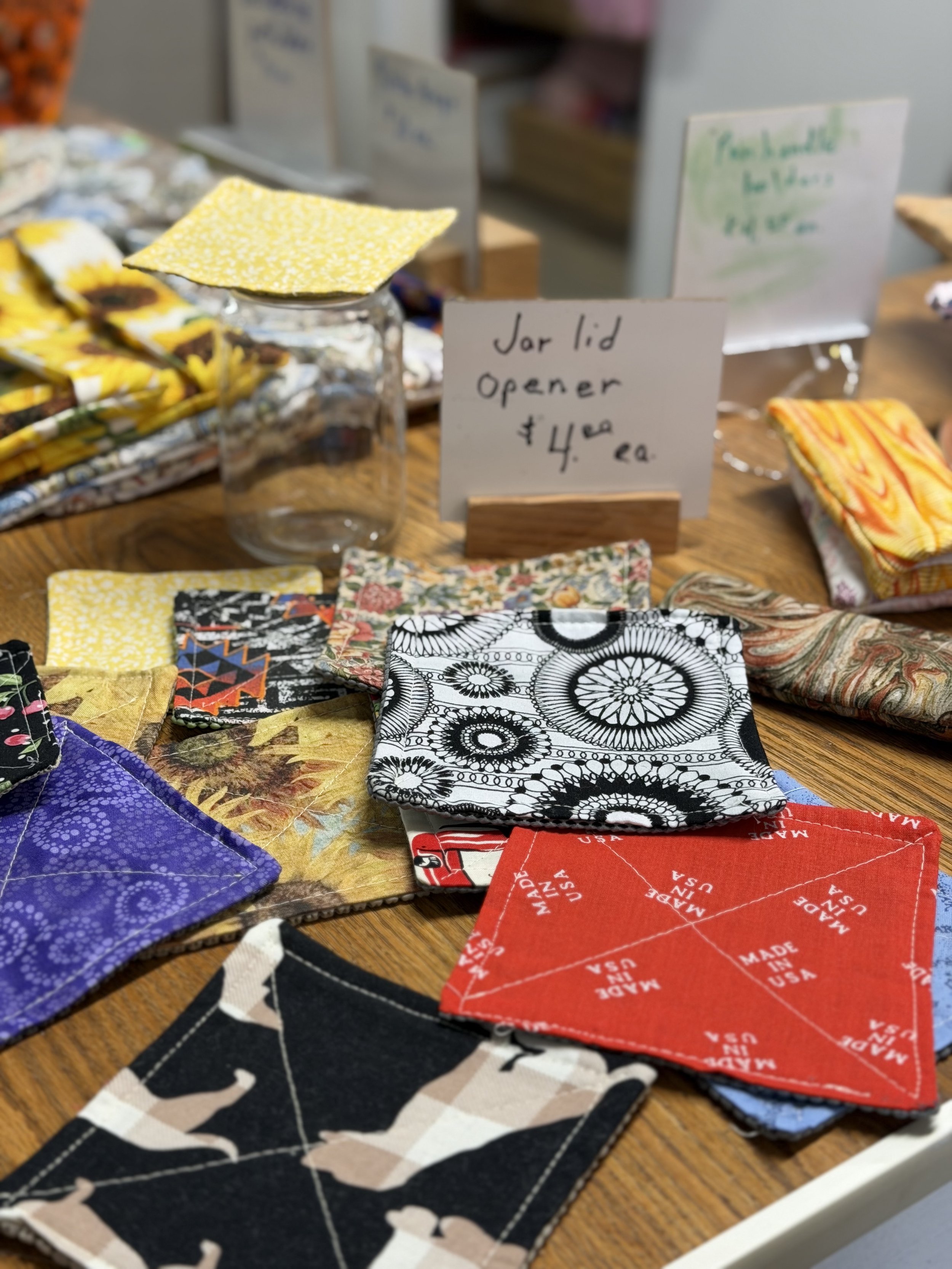 A display of fabric swatches and handmade fabric items on a wooden table, including a clear jar with a yellow cloth on top. A sign indicates 'Jar lid opener $4 ea'.
